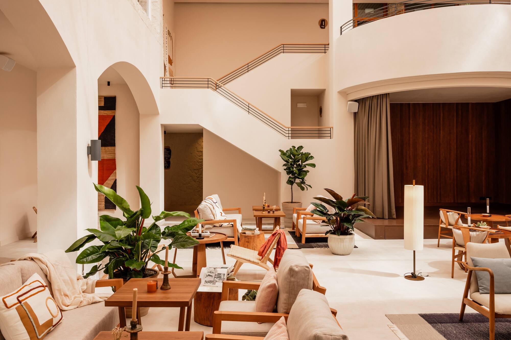 Modern hotel lobby lounge with wooden furniture, lush plants, and a grand staircase.