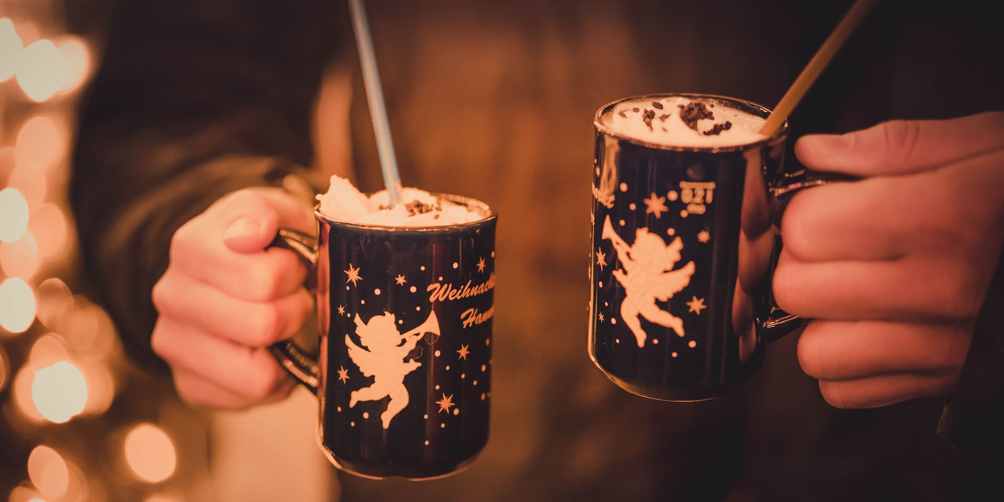 Hands hold dark Christmas mugs with angels and whipped cream, festive lights.