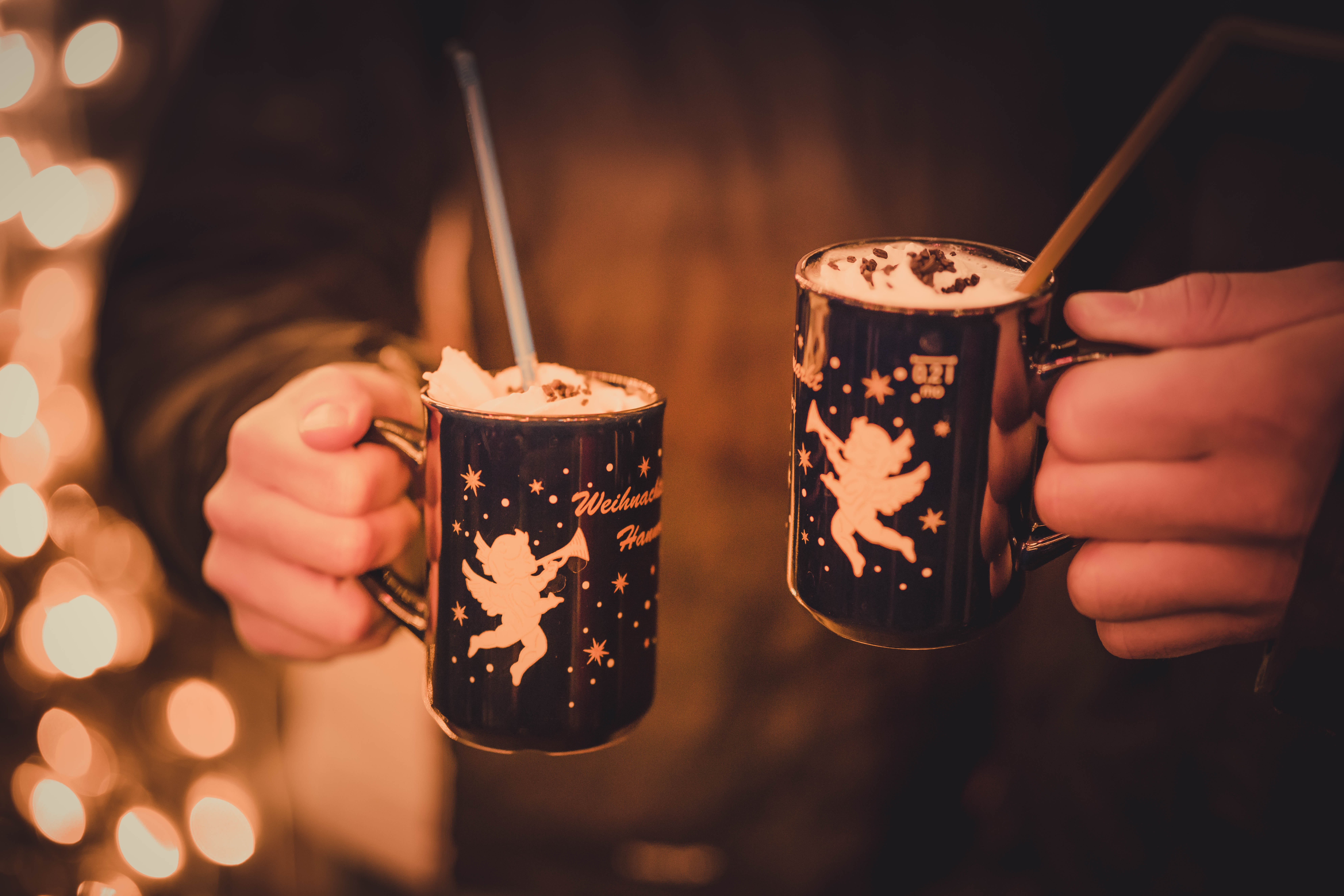 Hands hold dark Christmas mugs with angels and whipped cream, festive lights.