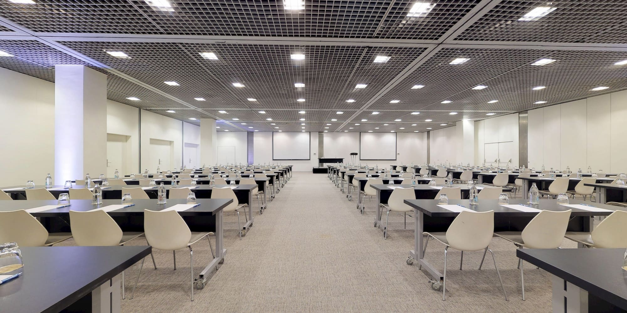 Spacious event venue with rows of tables, chairs, and presentation screens.