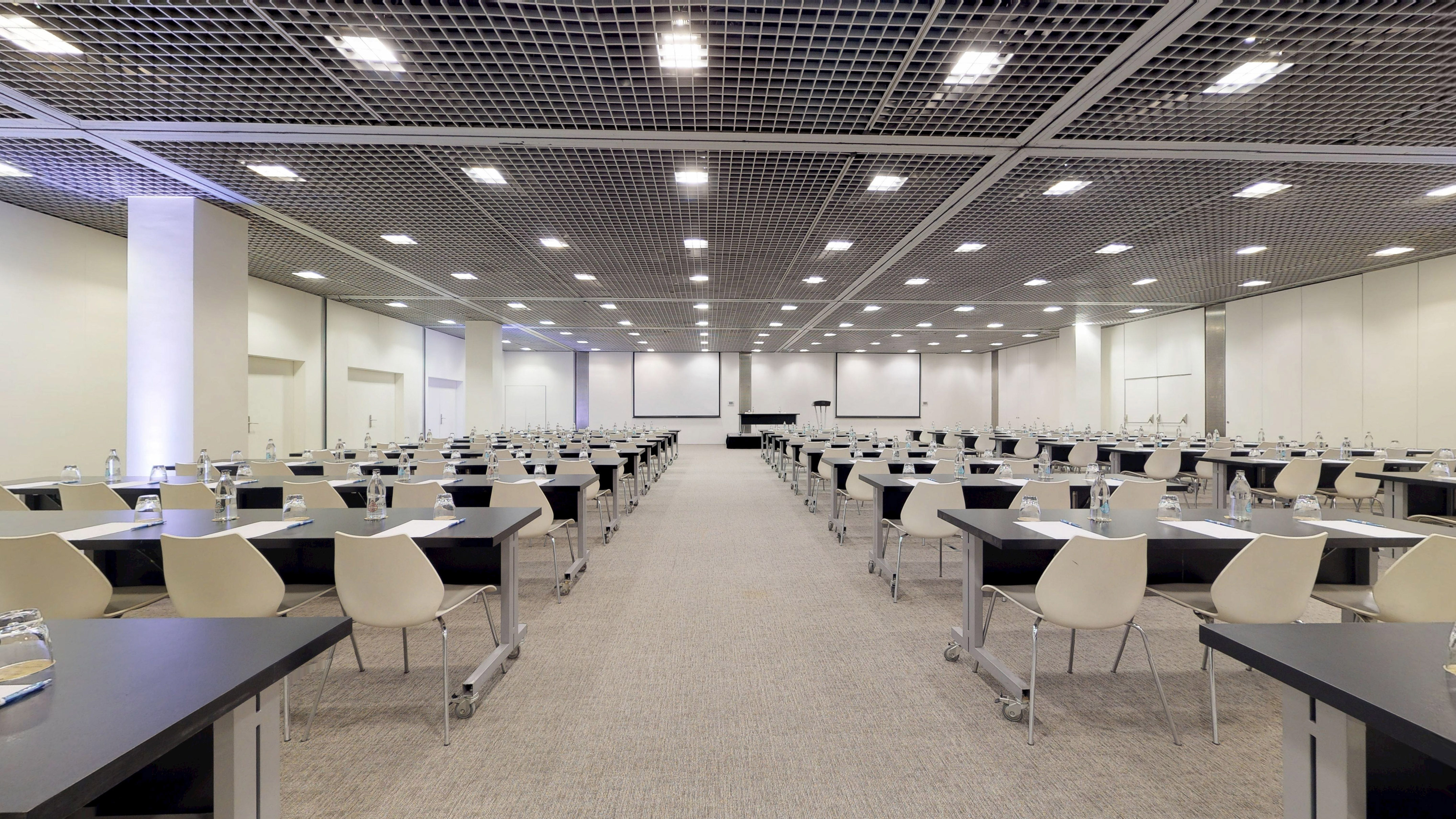 Spacious event venue with rows of tables, chairs, and presentation screens.