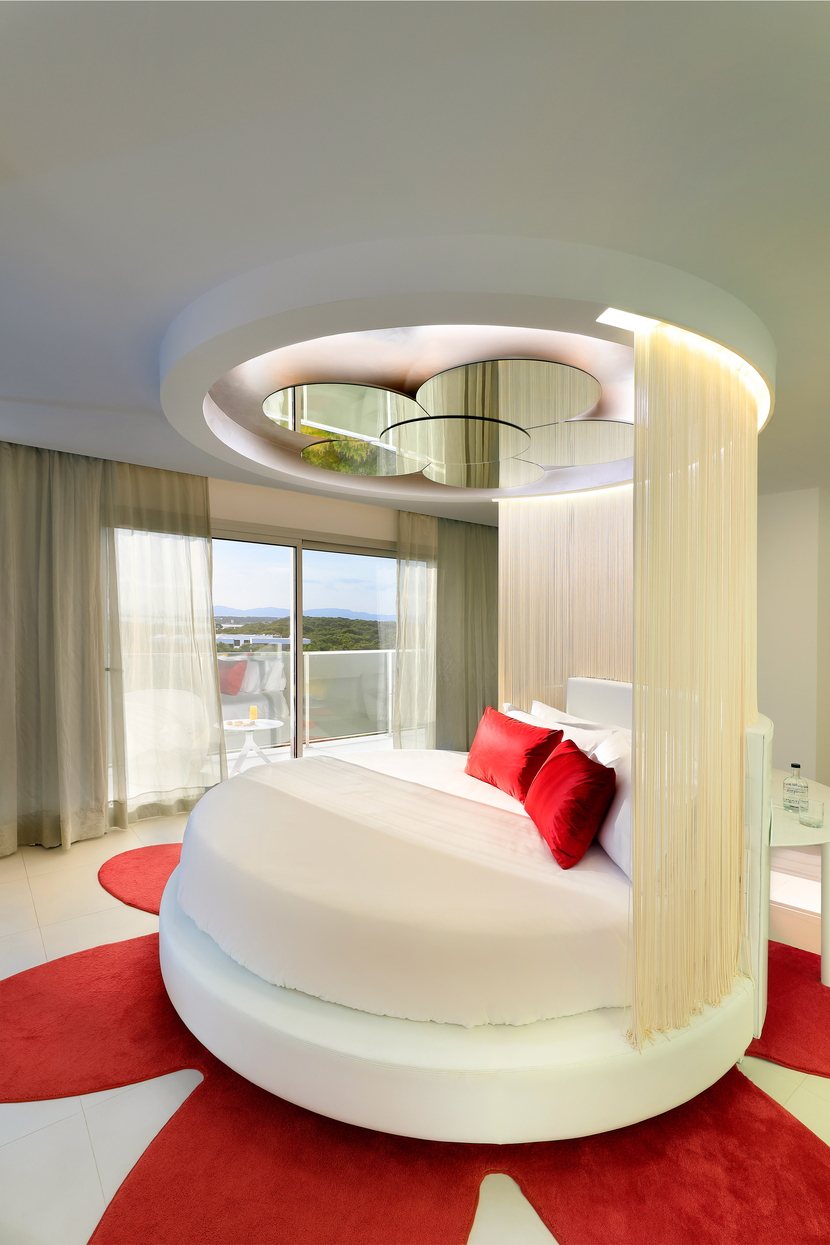 Modern luxury bedroom with round bed, red accents, balcony, and scenic view.