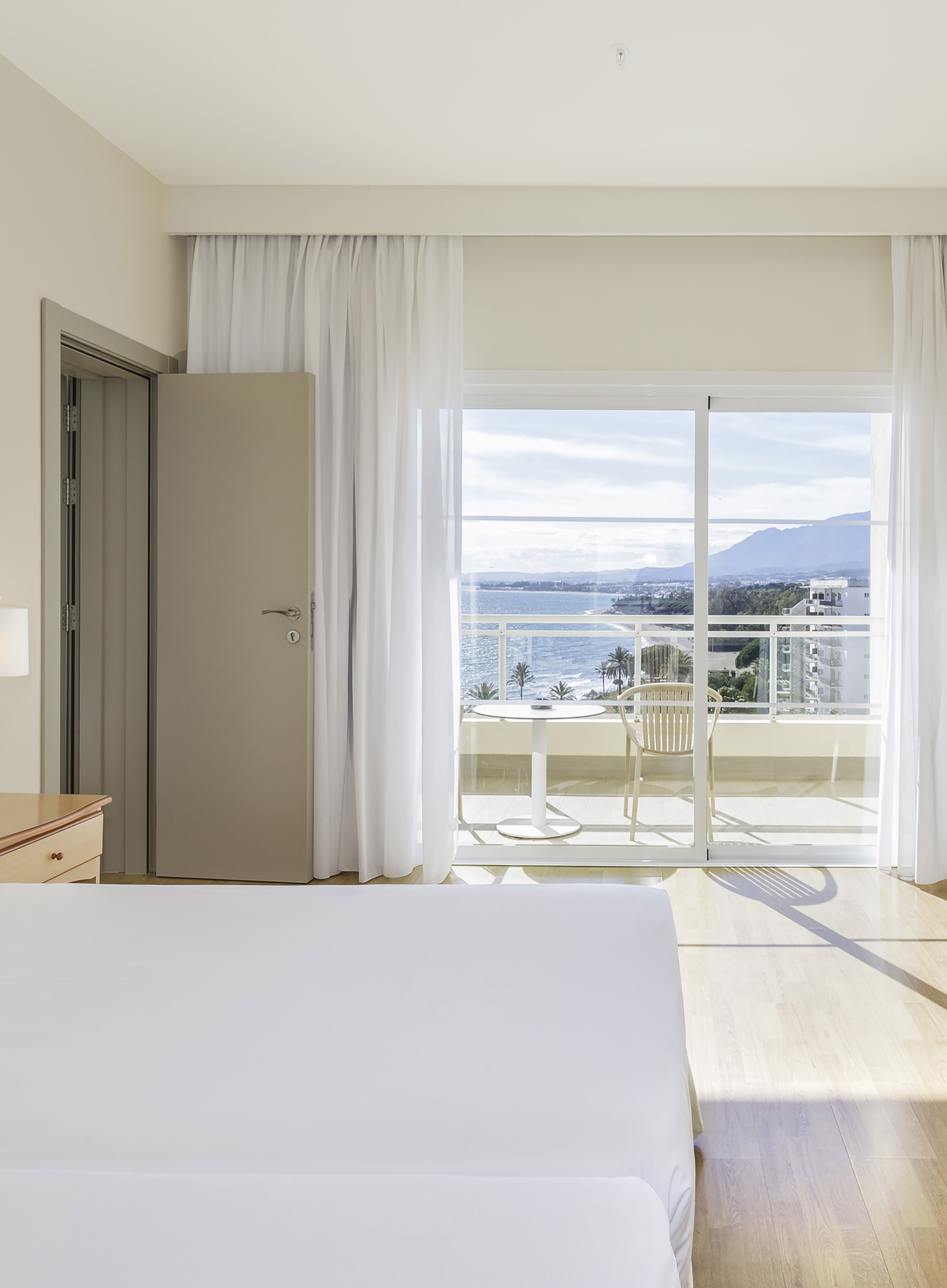 Bright hotel room with white bed, balcony, sea view, beach, and mountains.