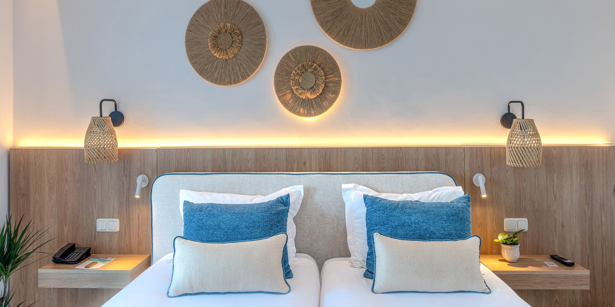 Hotel bedroom with twin beds, blue pillows, natural wood, and rattan decor.