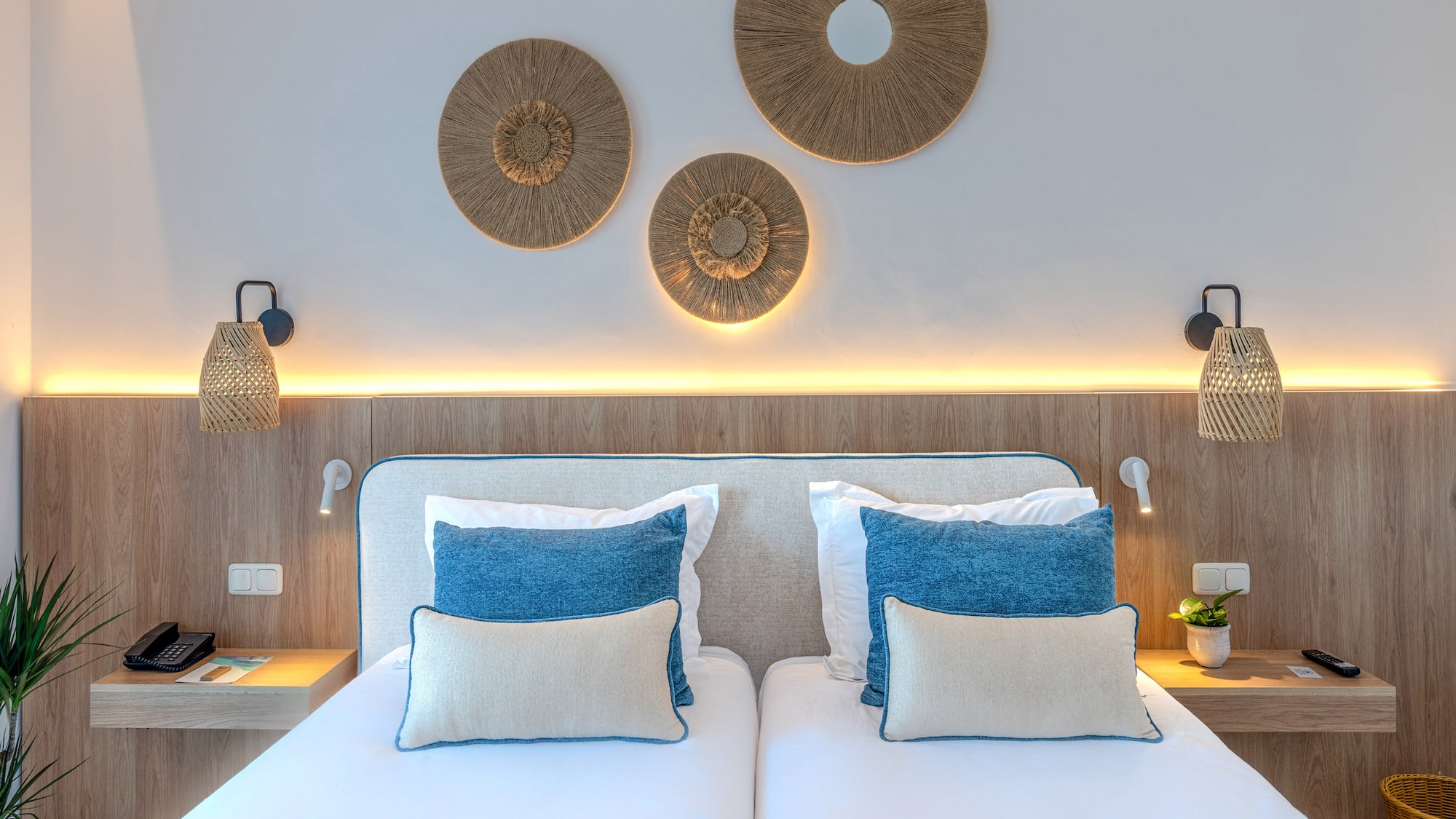 Hotel bedroom with twin beds, blue pillows, natural wood, and rattan decor.