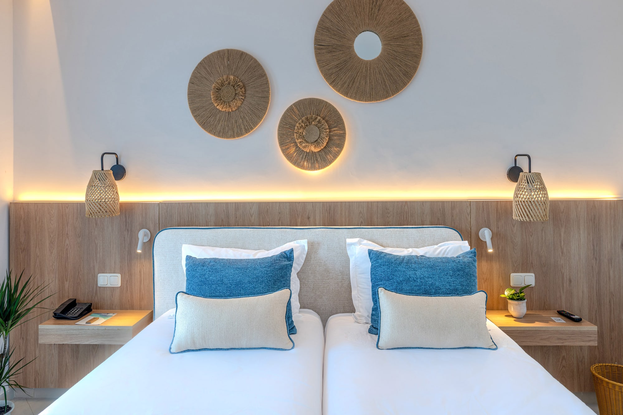 Hotel bedroom with twin beds, blue pillows, natural wood, and rattan decor.