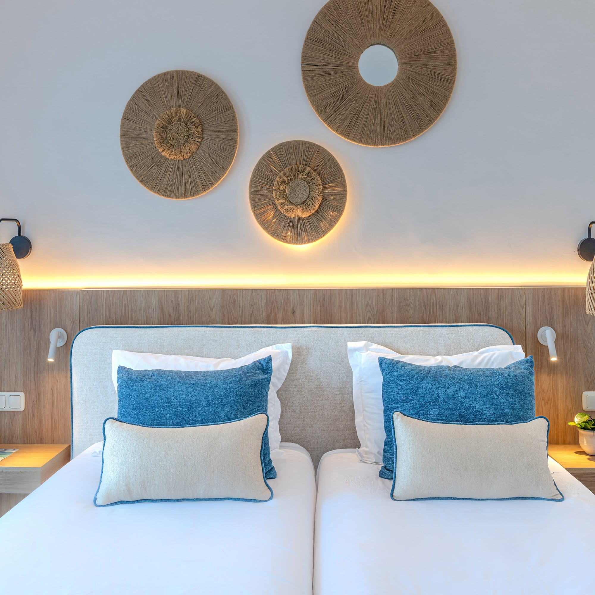 Hotel bedroom with twin beds, blue pillows, natural wood, and rattan decor.