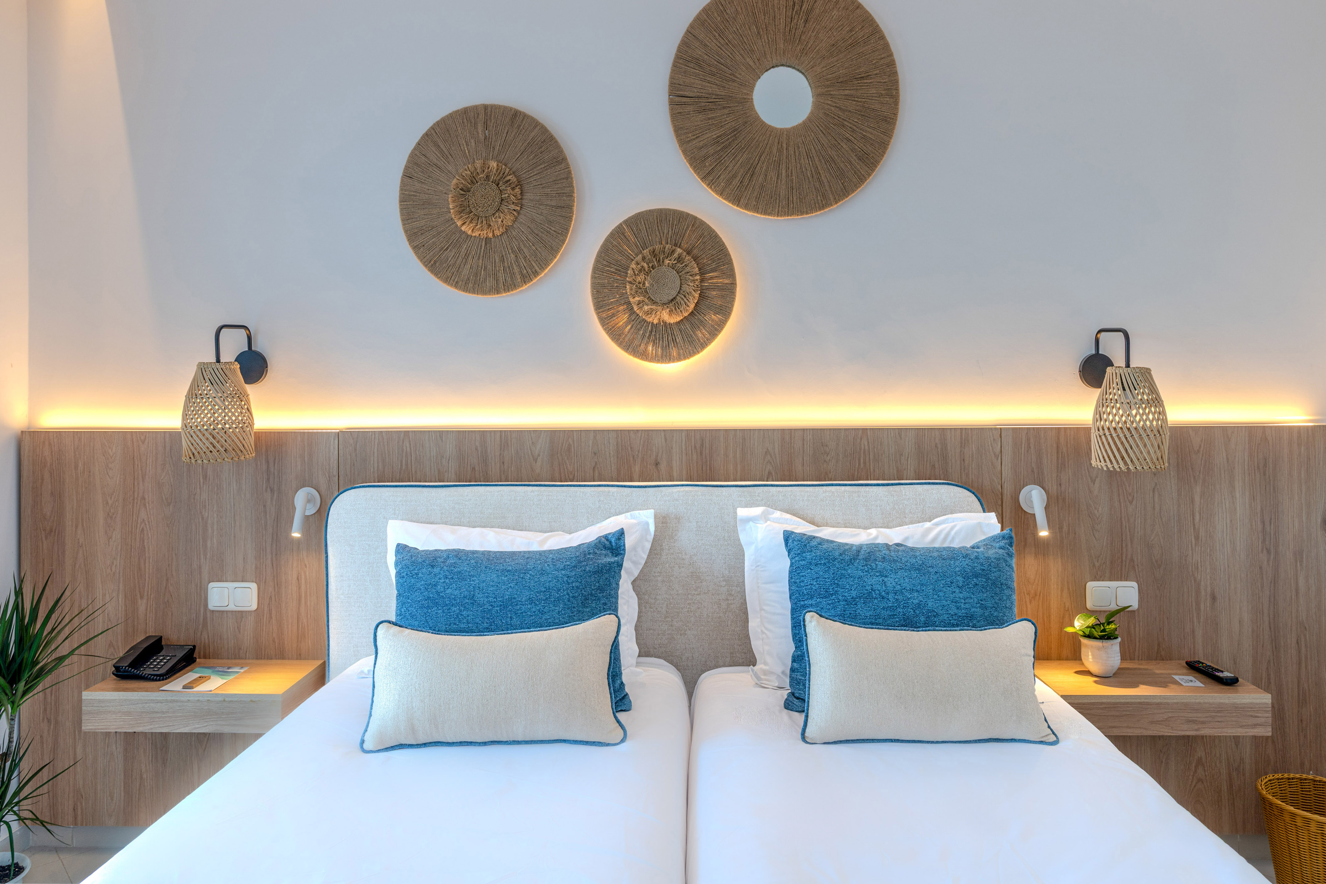 Hotel bedroom with twin beds, blue pillows, natural wood, and rattan decor.