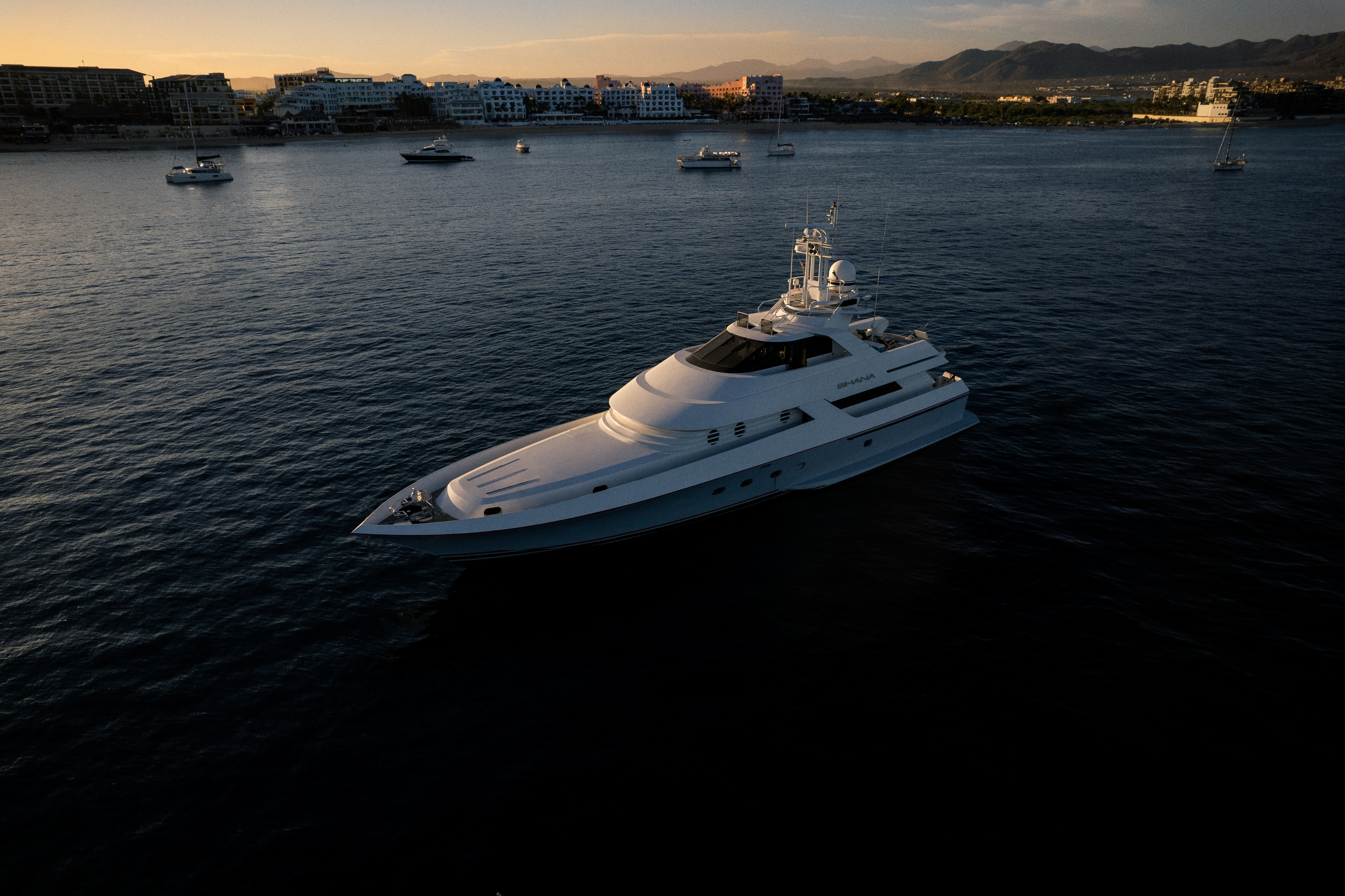 Luxury yacht 'SHANA' in bay at sunset with coastal resort.