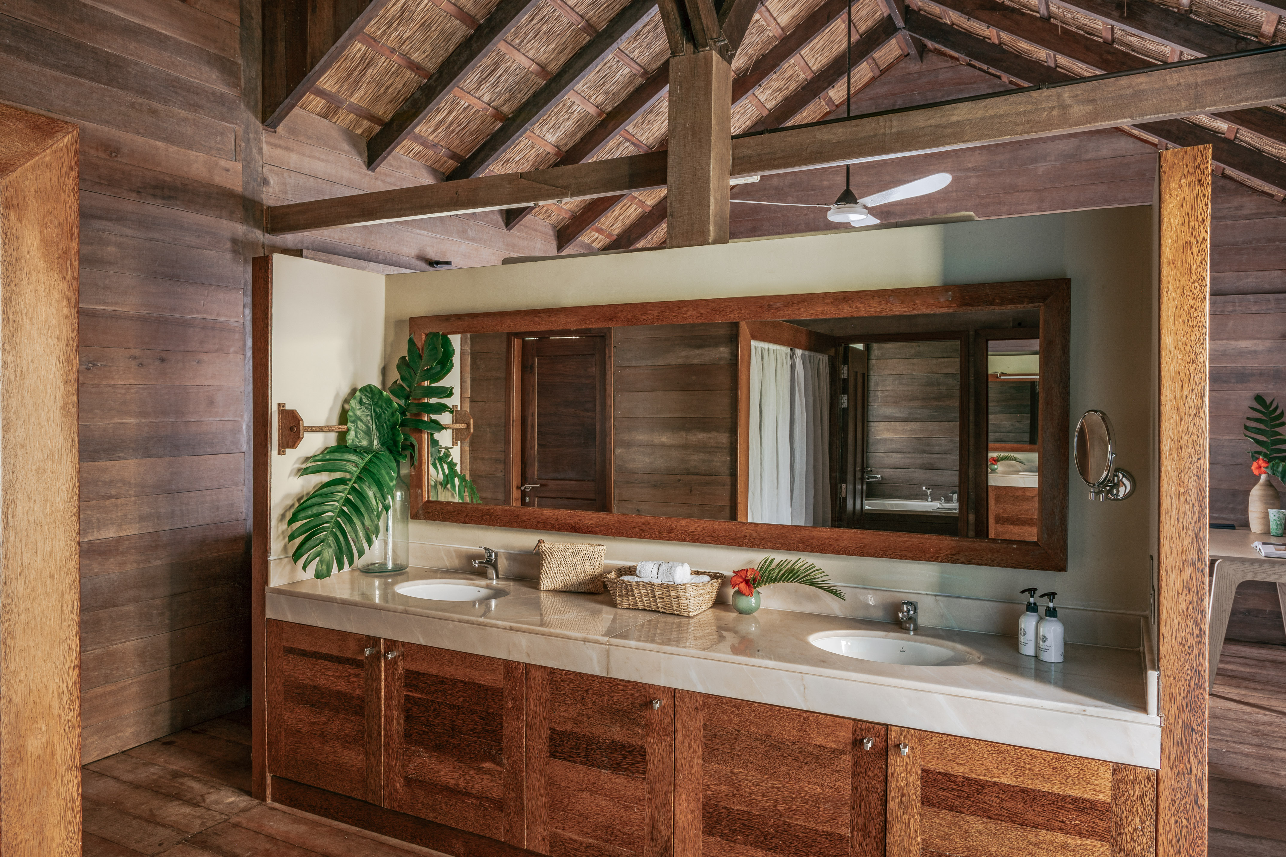 Tropical wooden bathroom with double sink vanity, mirror, and thatched ceiling.