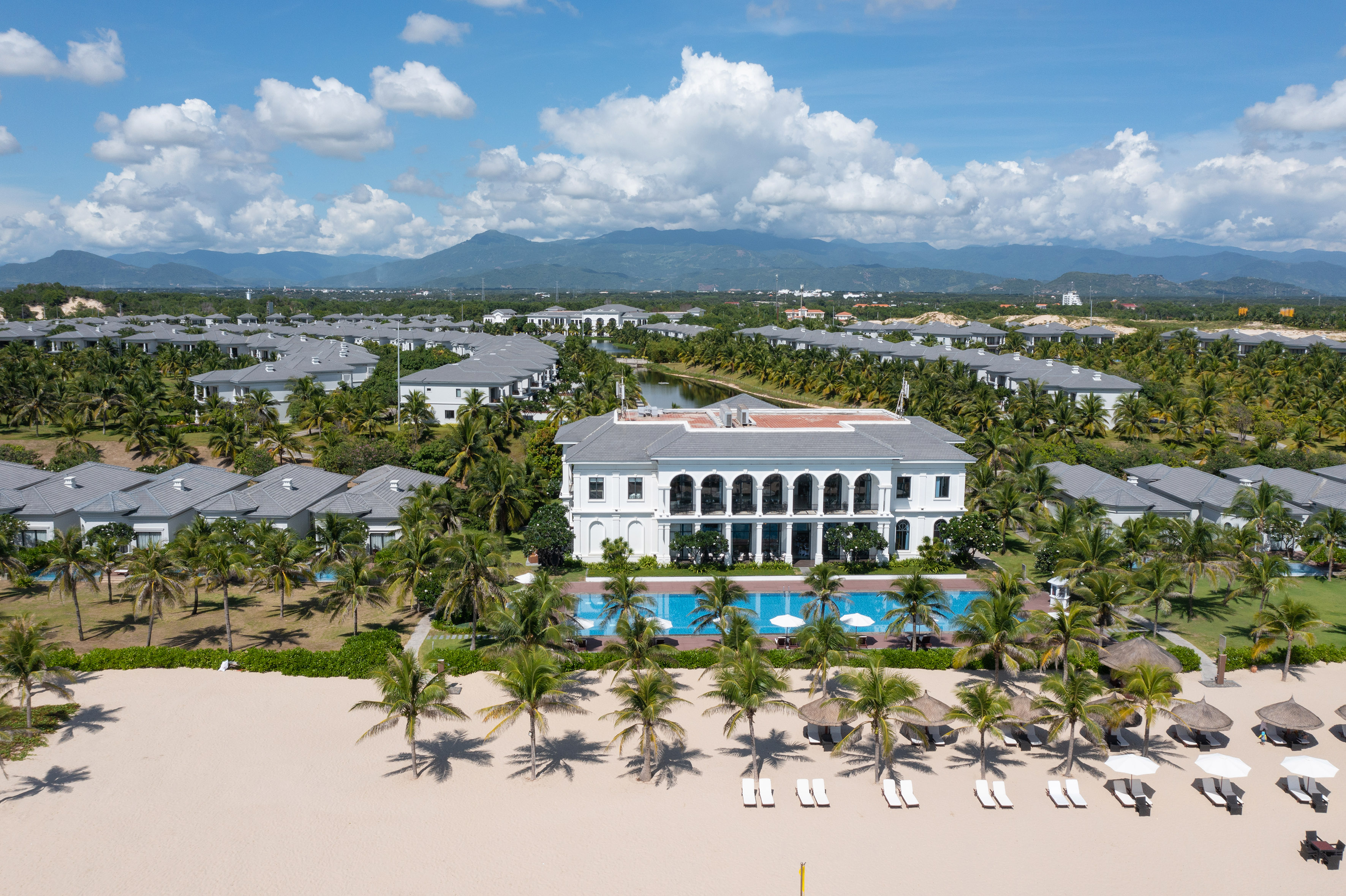 Luxury beachfront resort with white villas, pool, palm trees, and mountains.