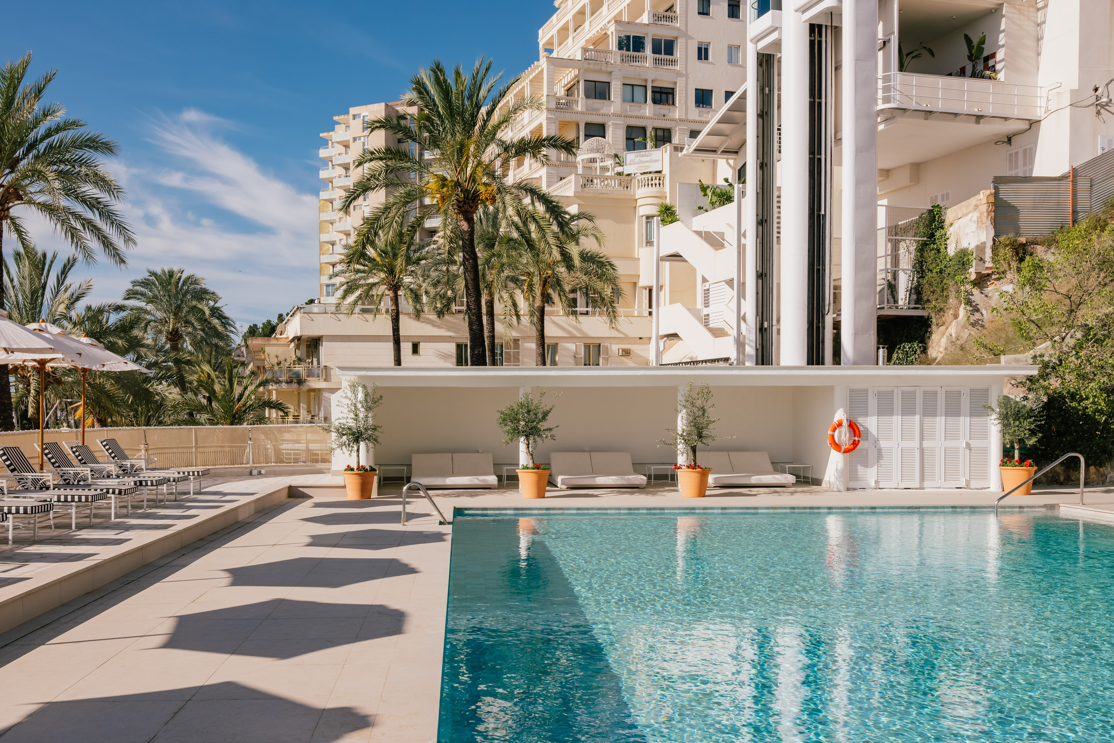 Mediterranean hotel pool with palm trees, loungers, white buildings.