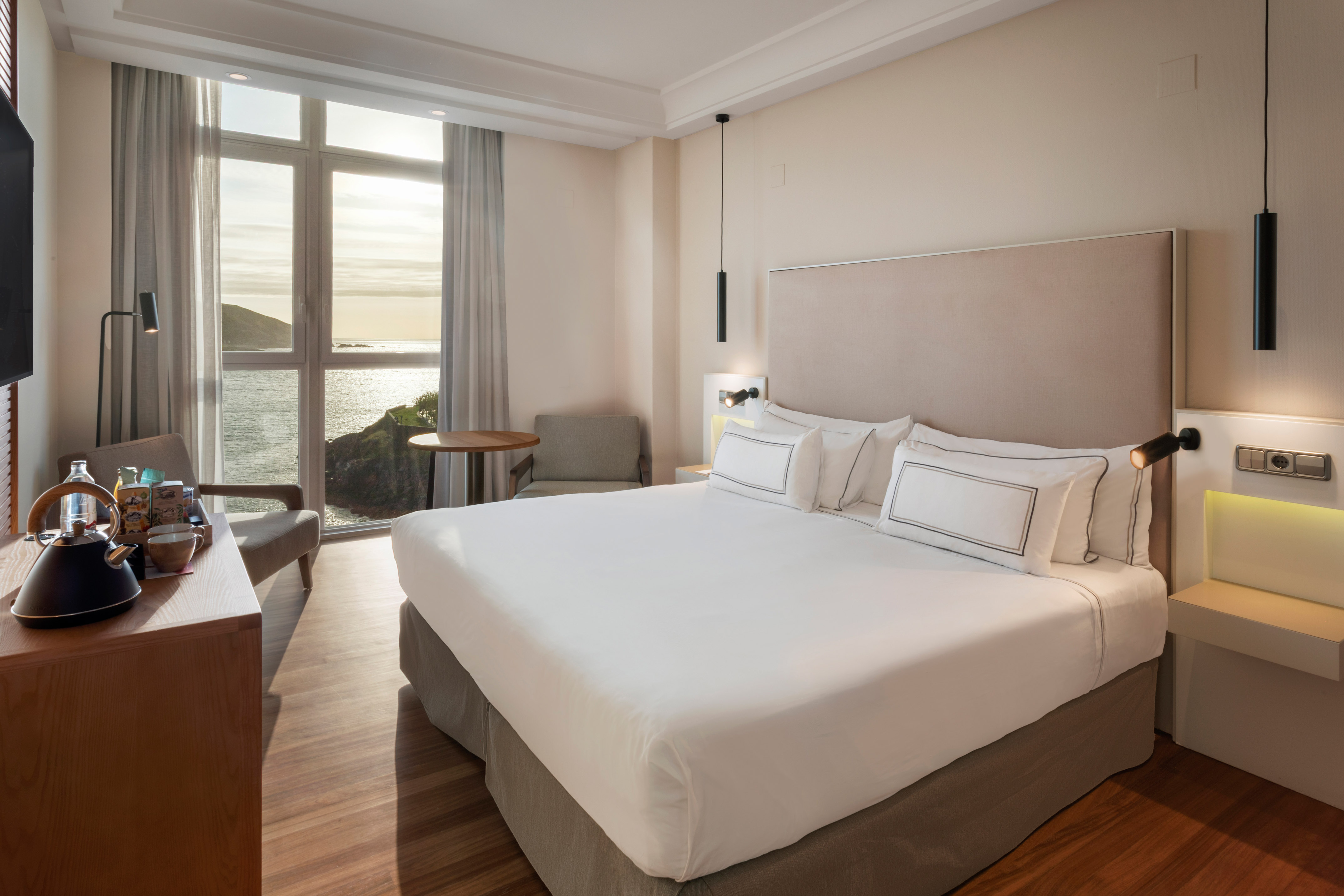 Modern hotel room with large bed, wooden floor, and ocean view from window.
