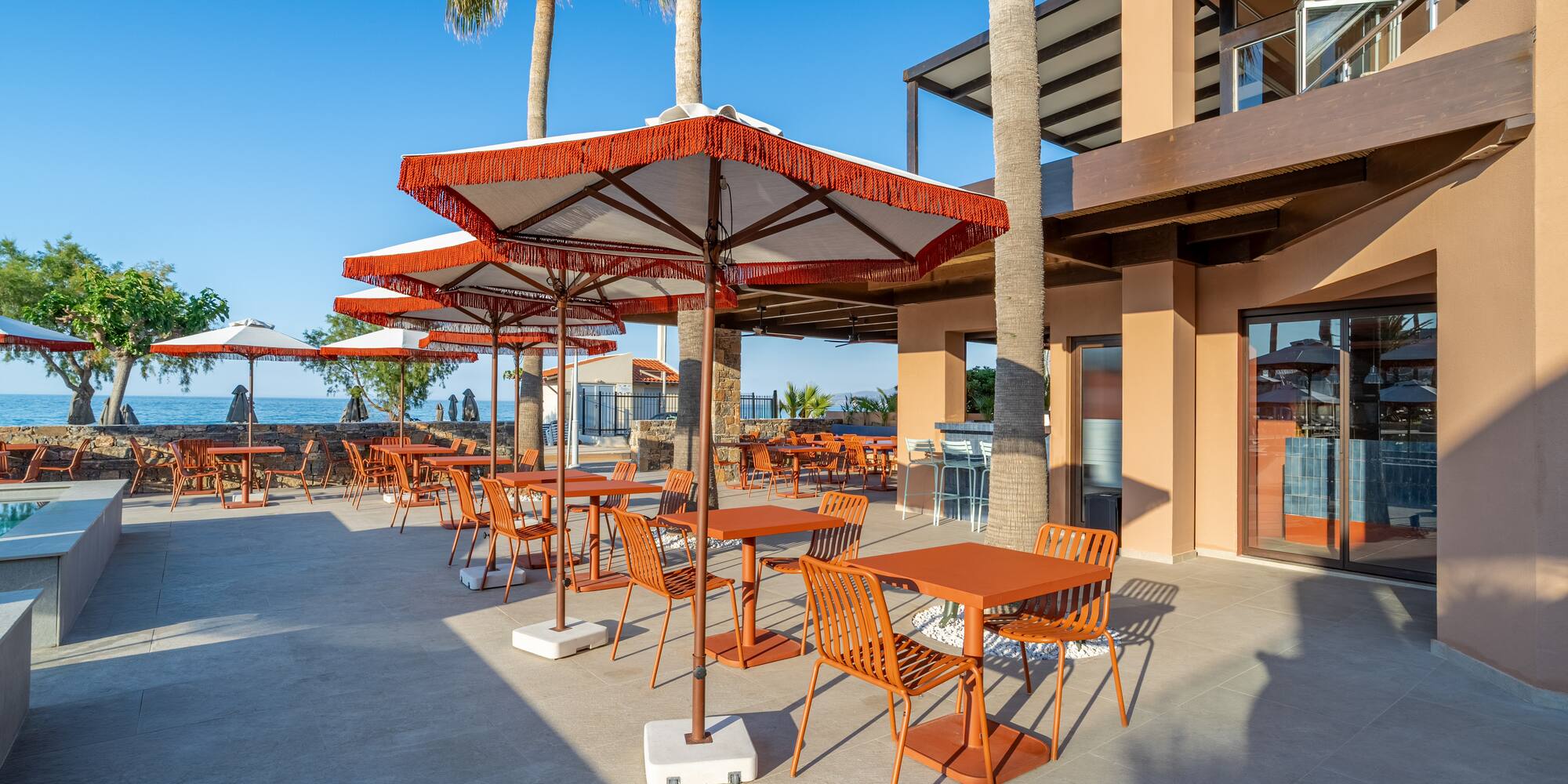 Outdoor dining patio with orange tables, umbrellas, palm trees, sea view.