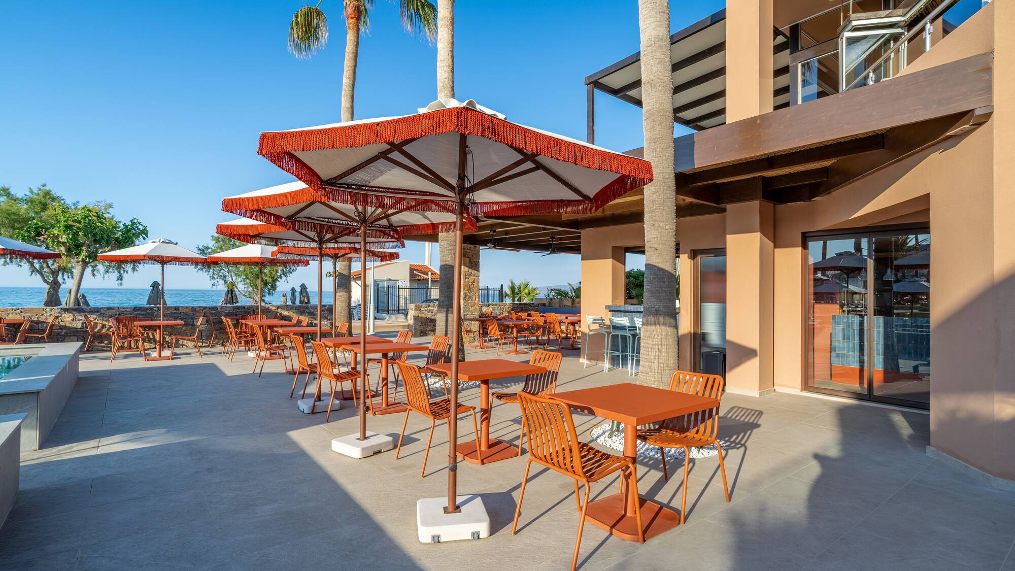 Outdoor dining patio with orange tables, umbrellas, palm trees, sea view.