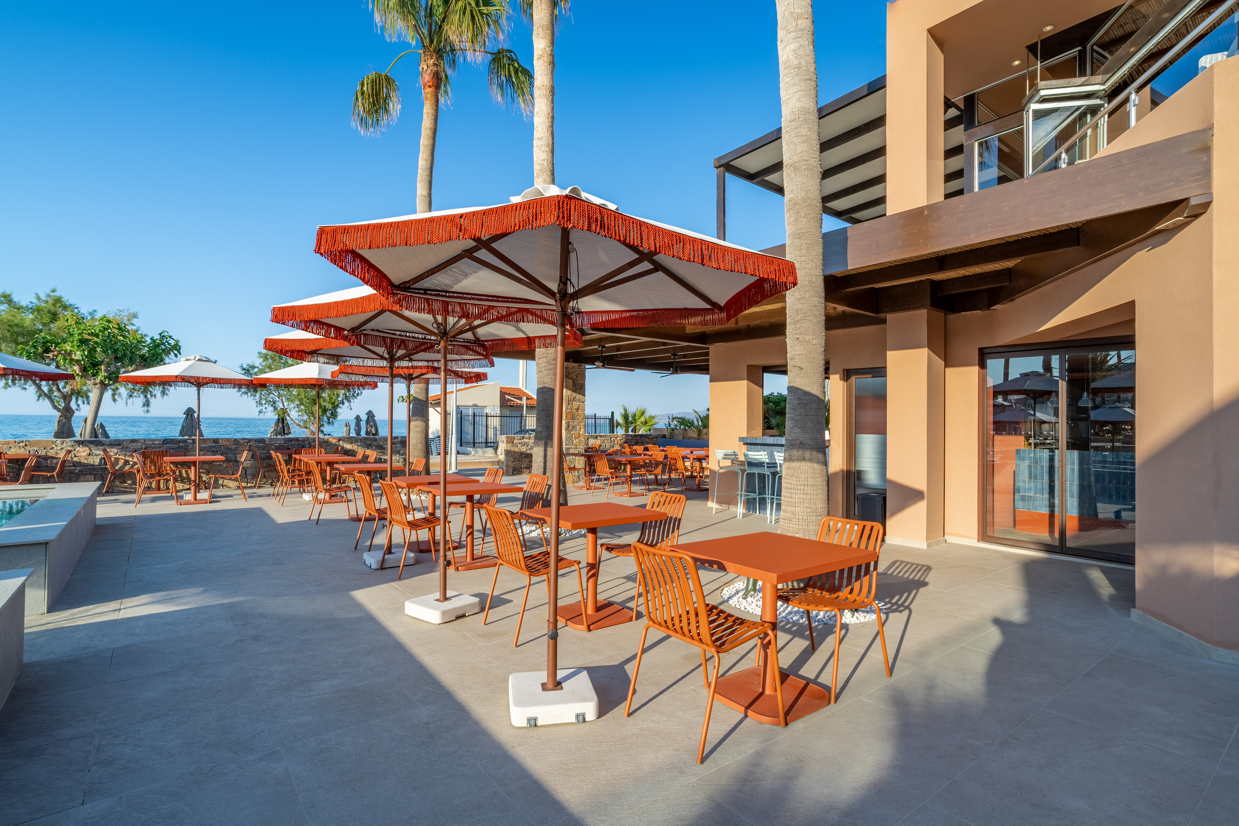 Outdoor dining patio with orange tables, umbrellas, palm trees, sea view.