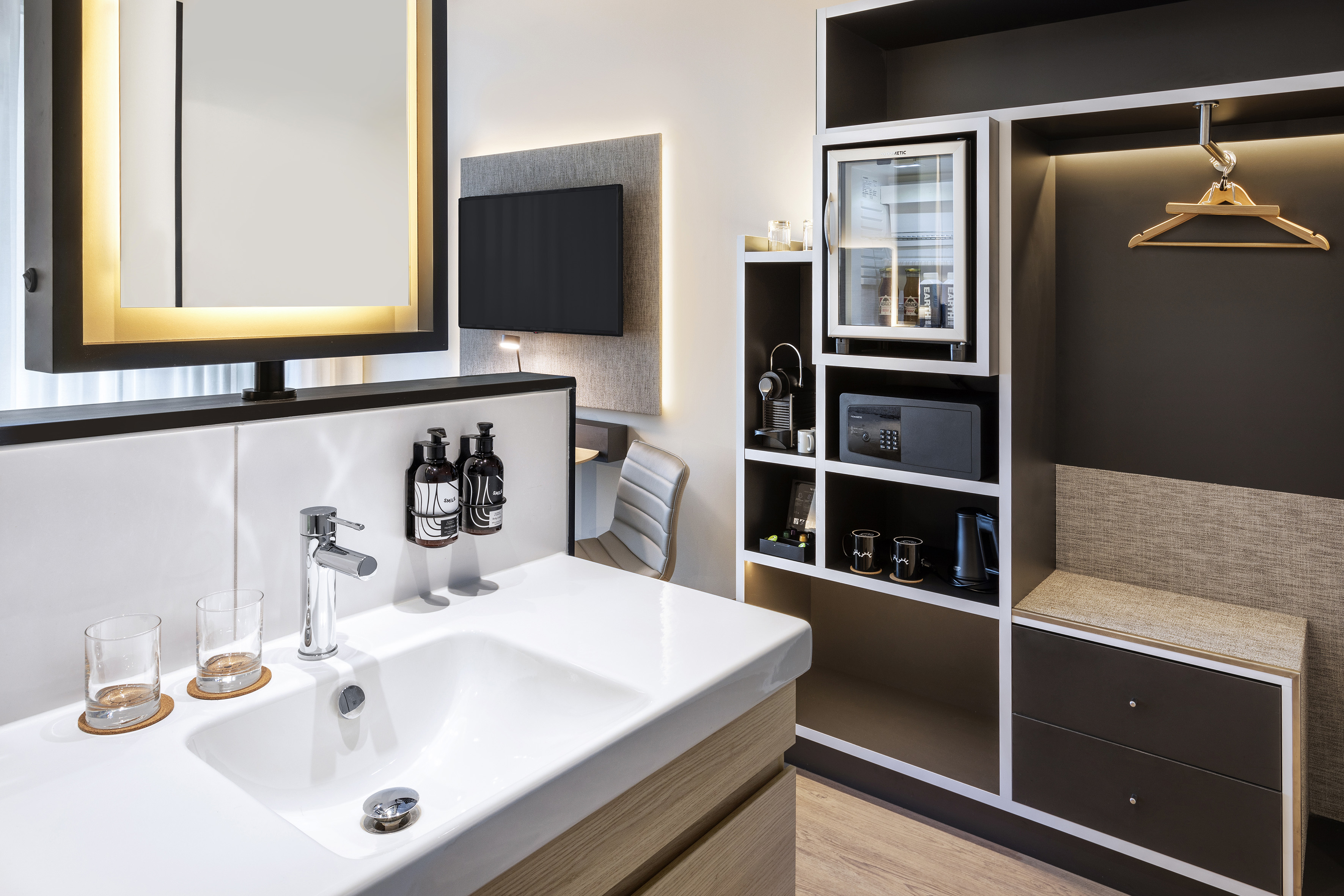 Modern hotel room with vanity, backlit mirror, TV, and integrated minibar.
