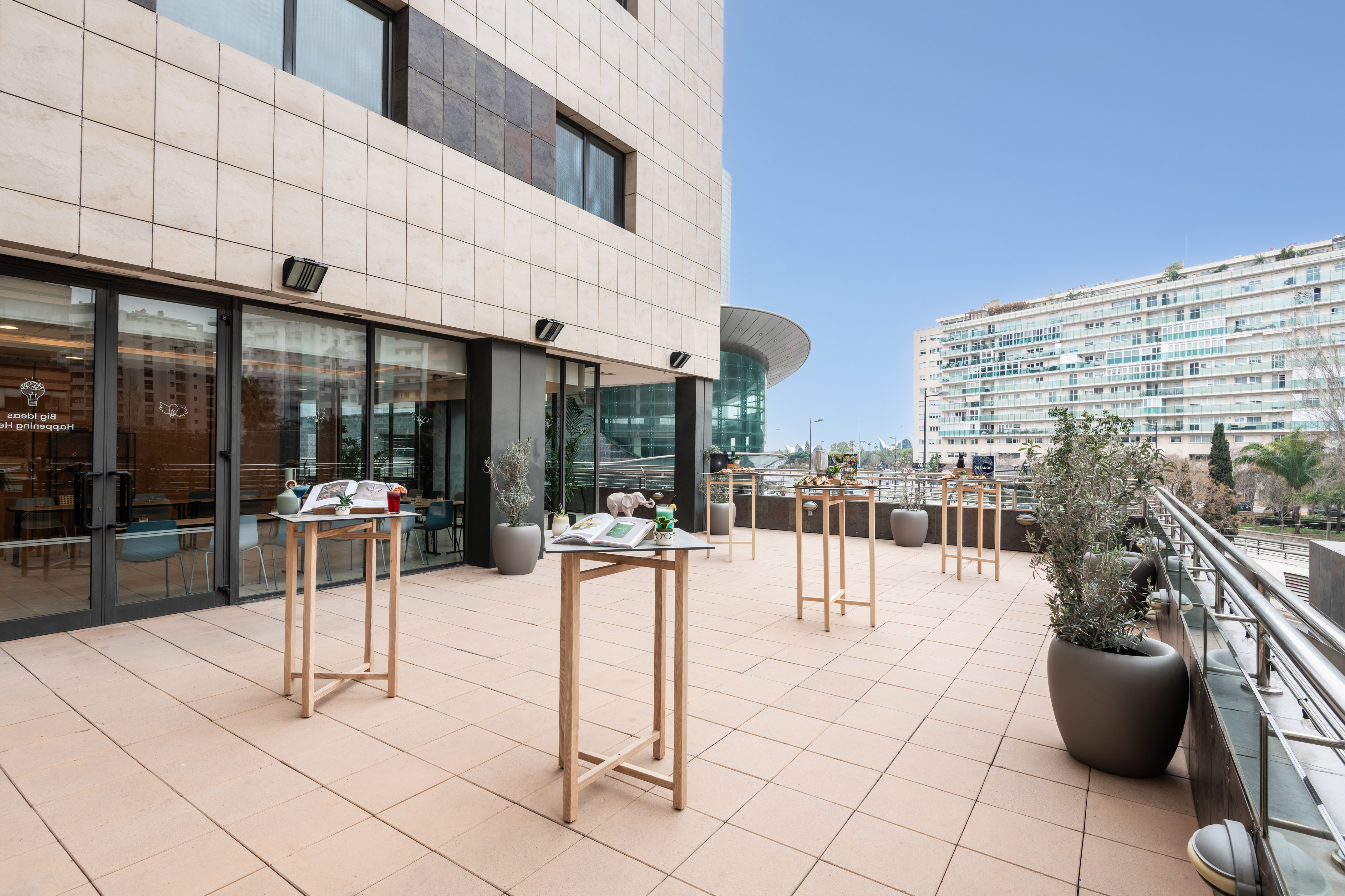Modern building terrace with cocktail tables, potted plants, and city background.