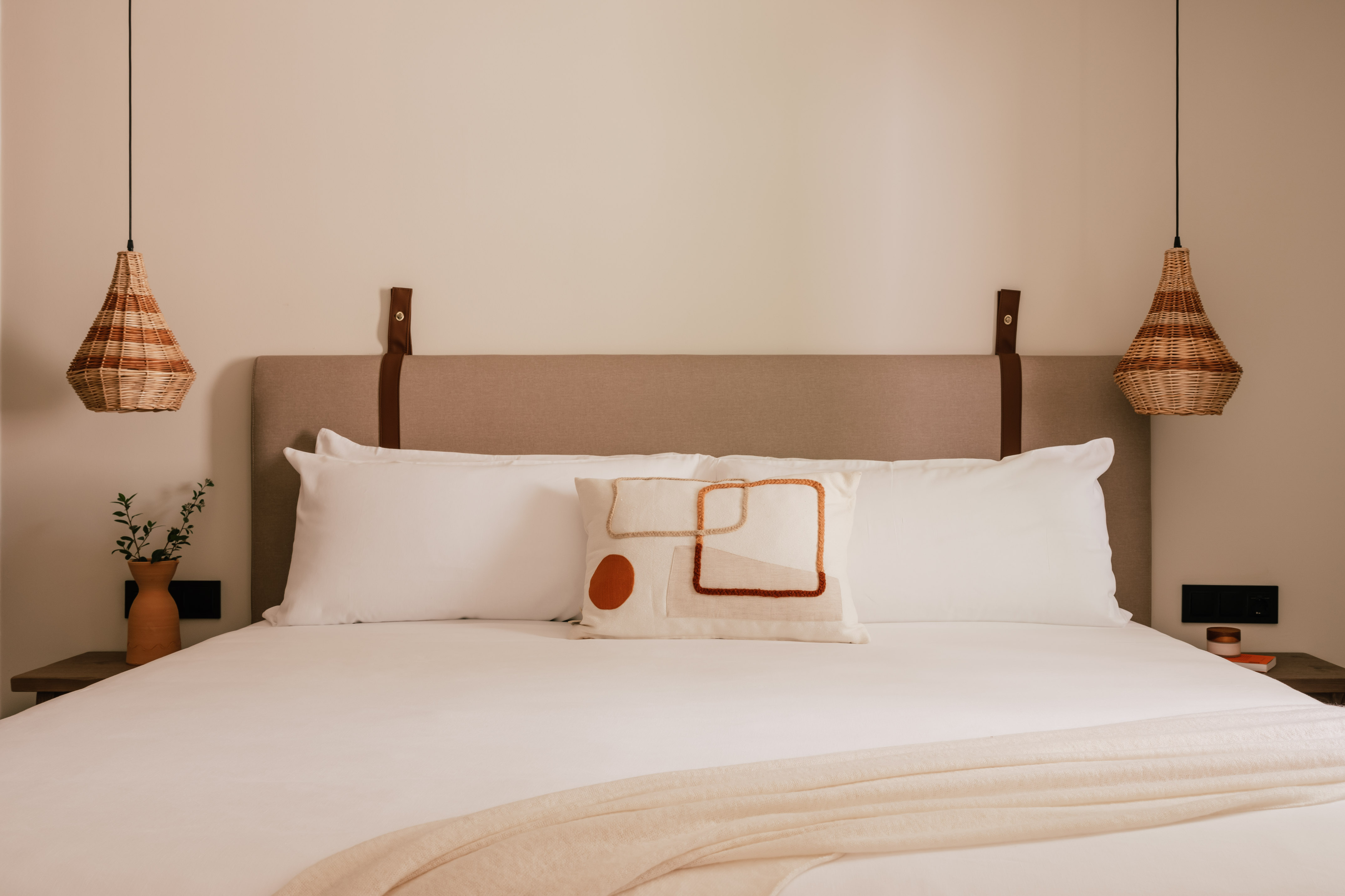 White hotel bed with abstract pillow, woven lights, upholstered headboard.