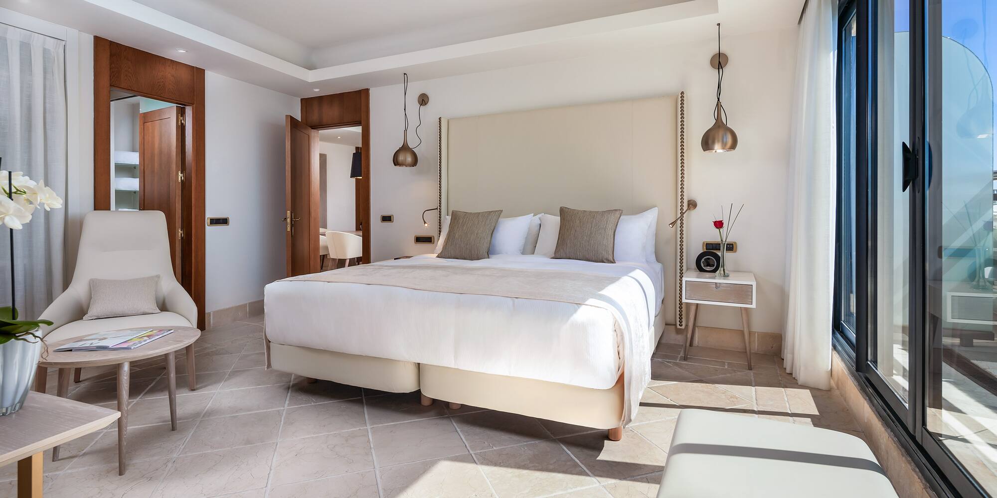 Modern luxury hotel bedroom with large bed, neutral decor, tiled floor, and balcony access.