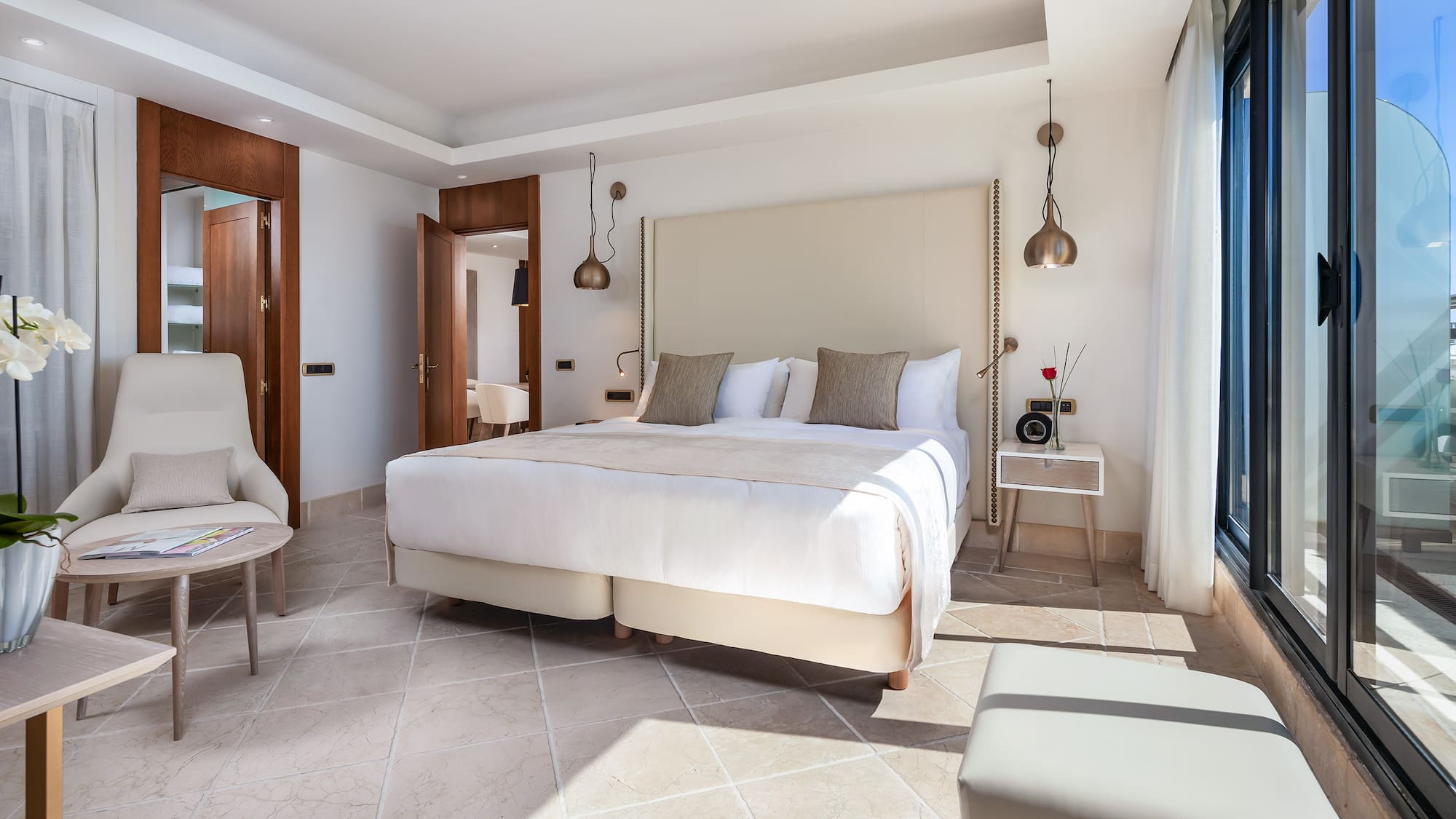 Modern luxury hotel bedroom with large bed, neutral decor, tiled floor, and balcony access.