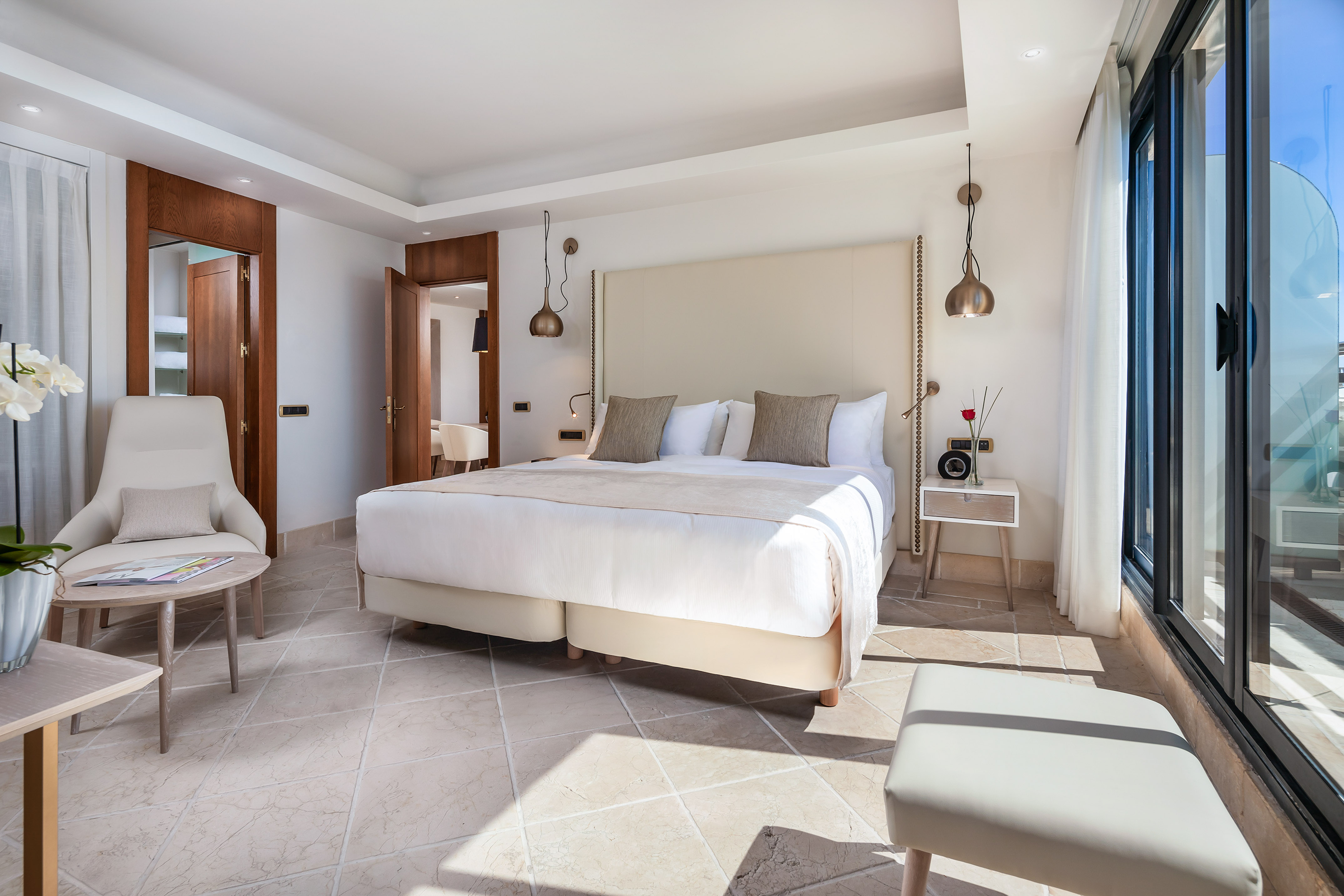 Modern luxury hotel bedroom with large bed, neutral decor, tiled floor, and balcony access.