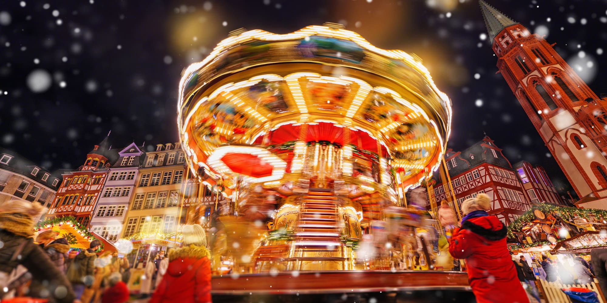 Illuminated carousel at a snowy Christmas market with historic buildings at night.