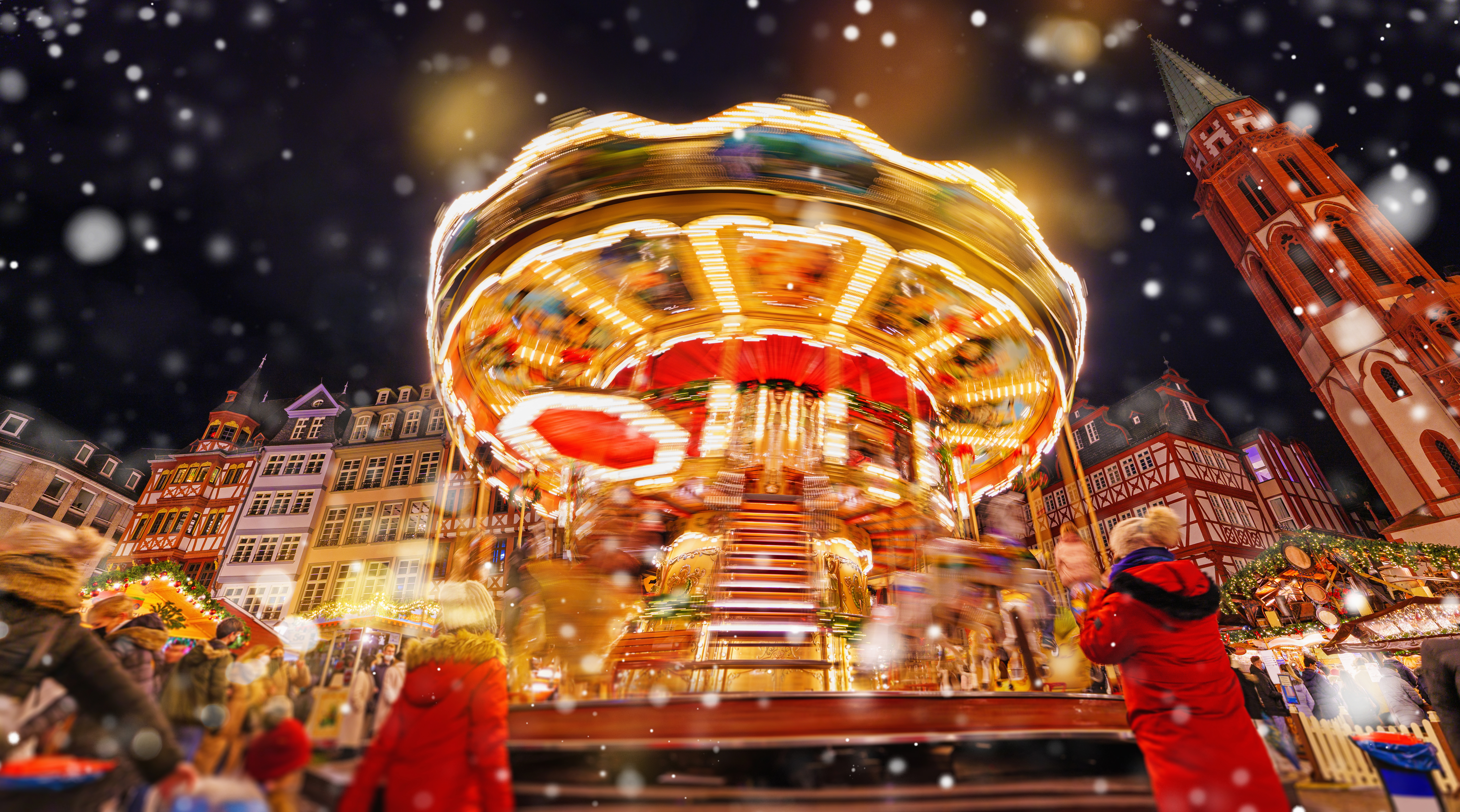 Illuminated carousel at a snowy Christmas market with historic buildings at night.