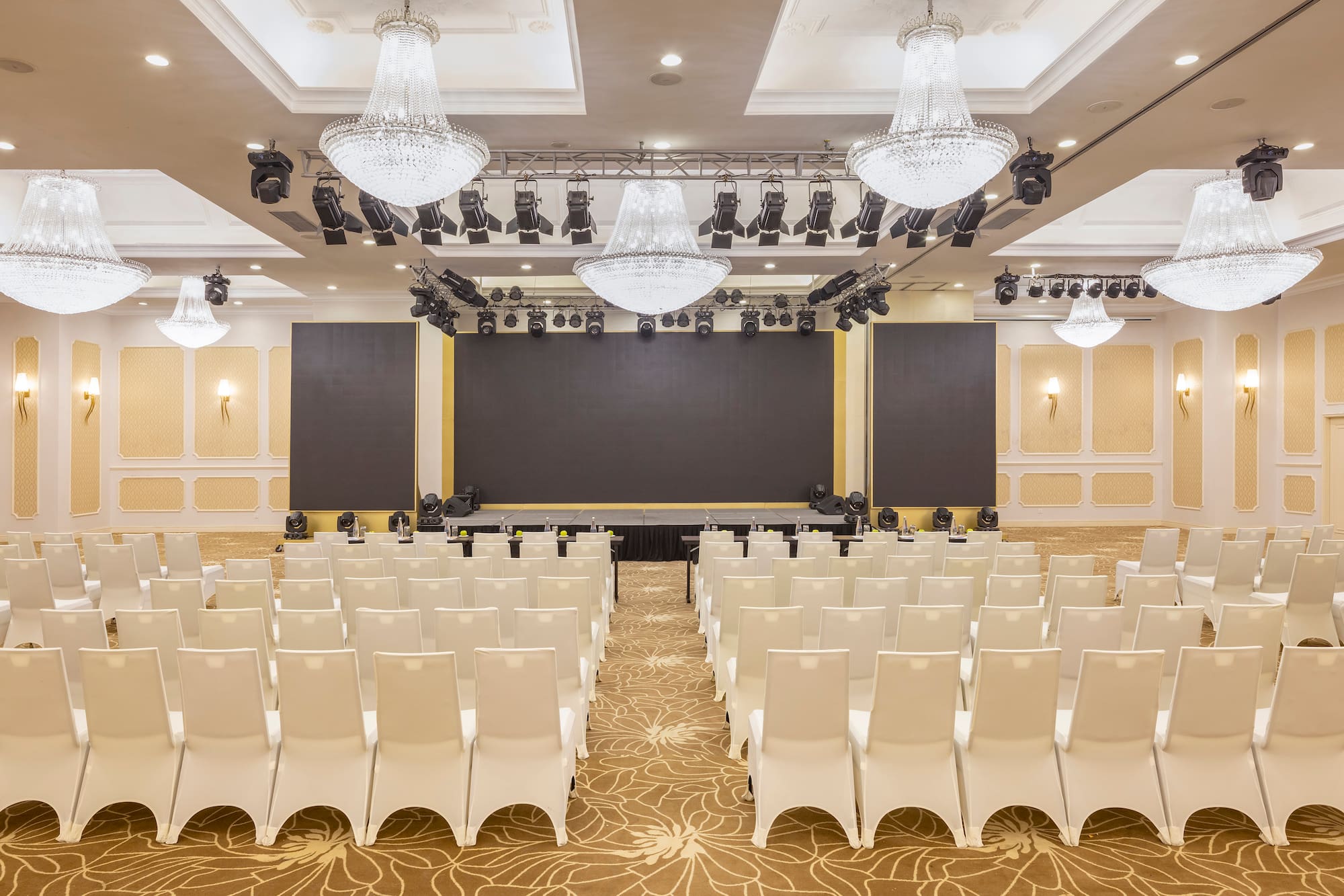 Empty elegant ballroom with white chairs, stage, screen, and chandeliers.