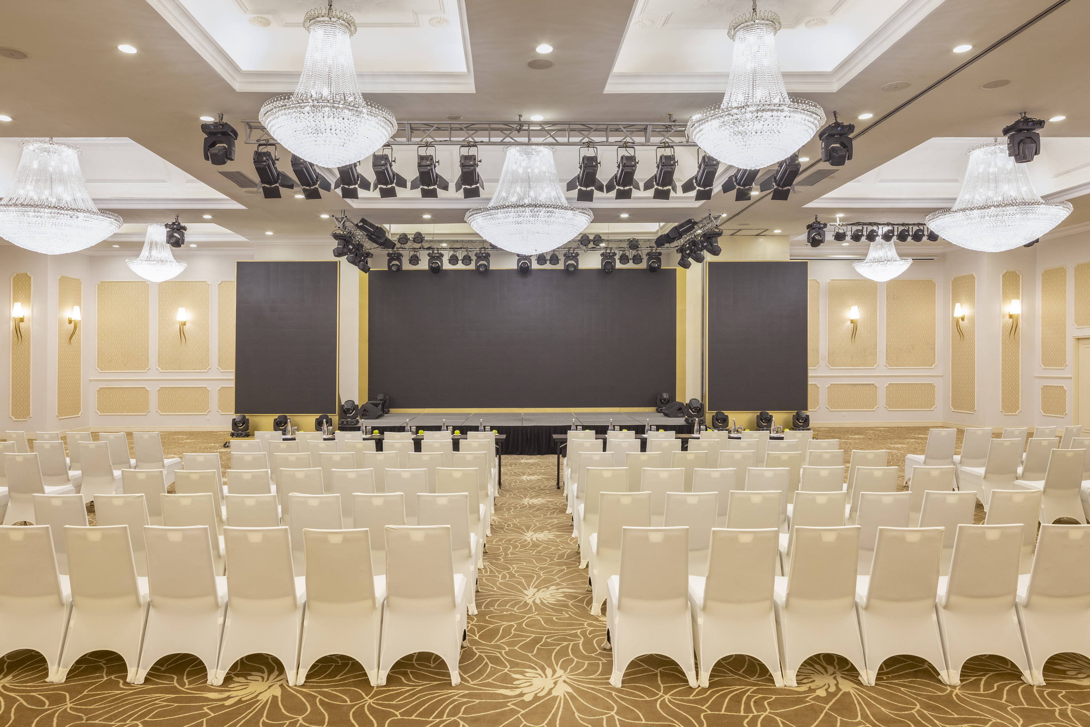 Empty elegant ballroom with white chairs, stage, screen, and chandeliers.