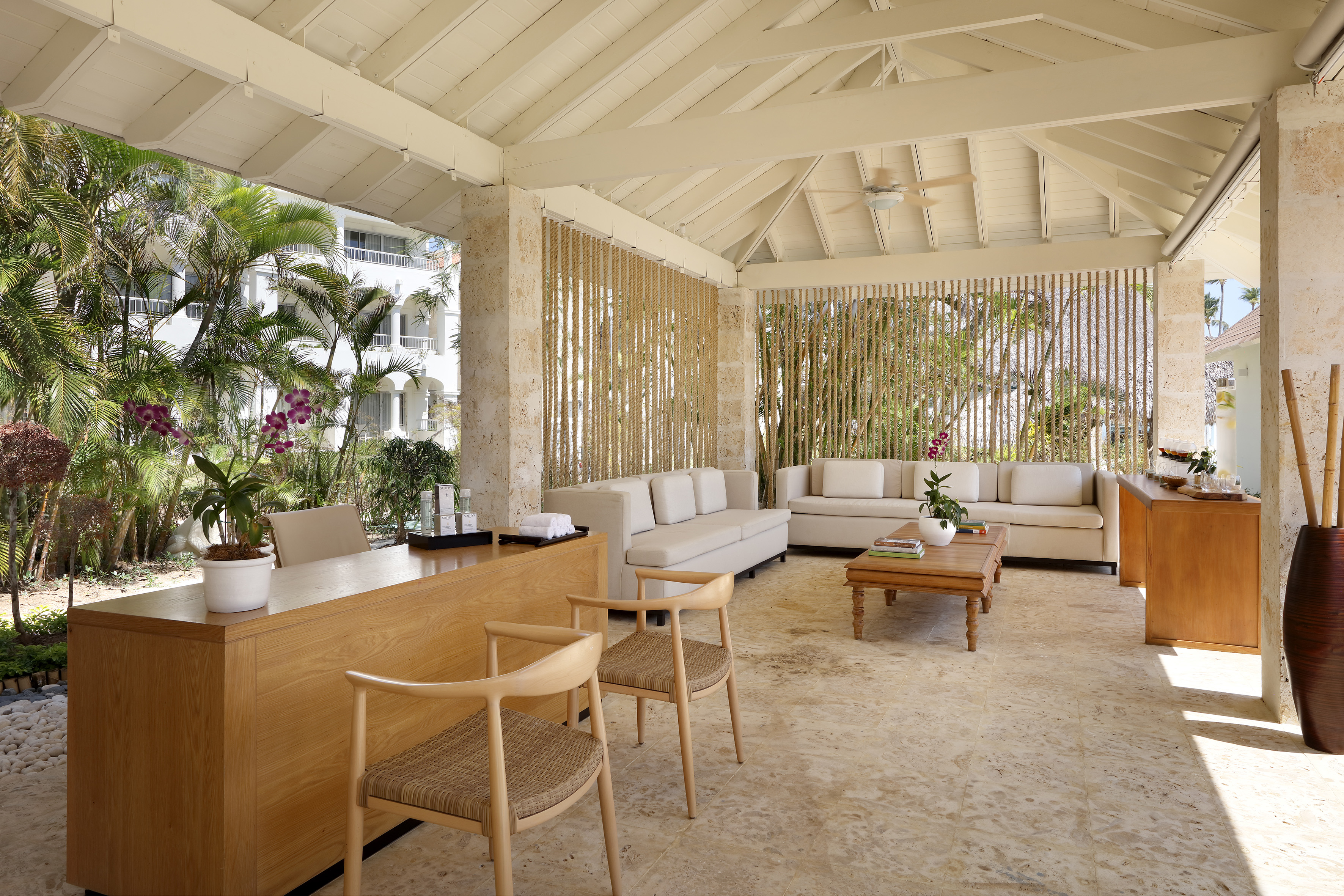 Outdoor tropical resort lobby with wooden desk, cream sofas, and palm trees.