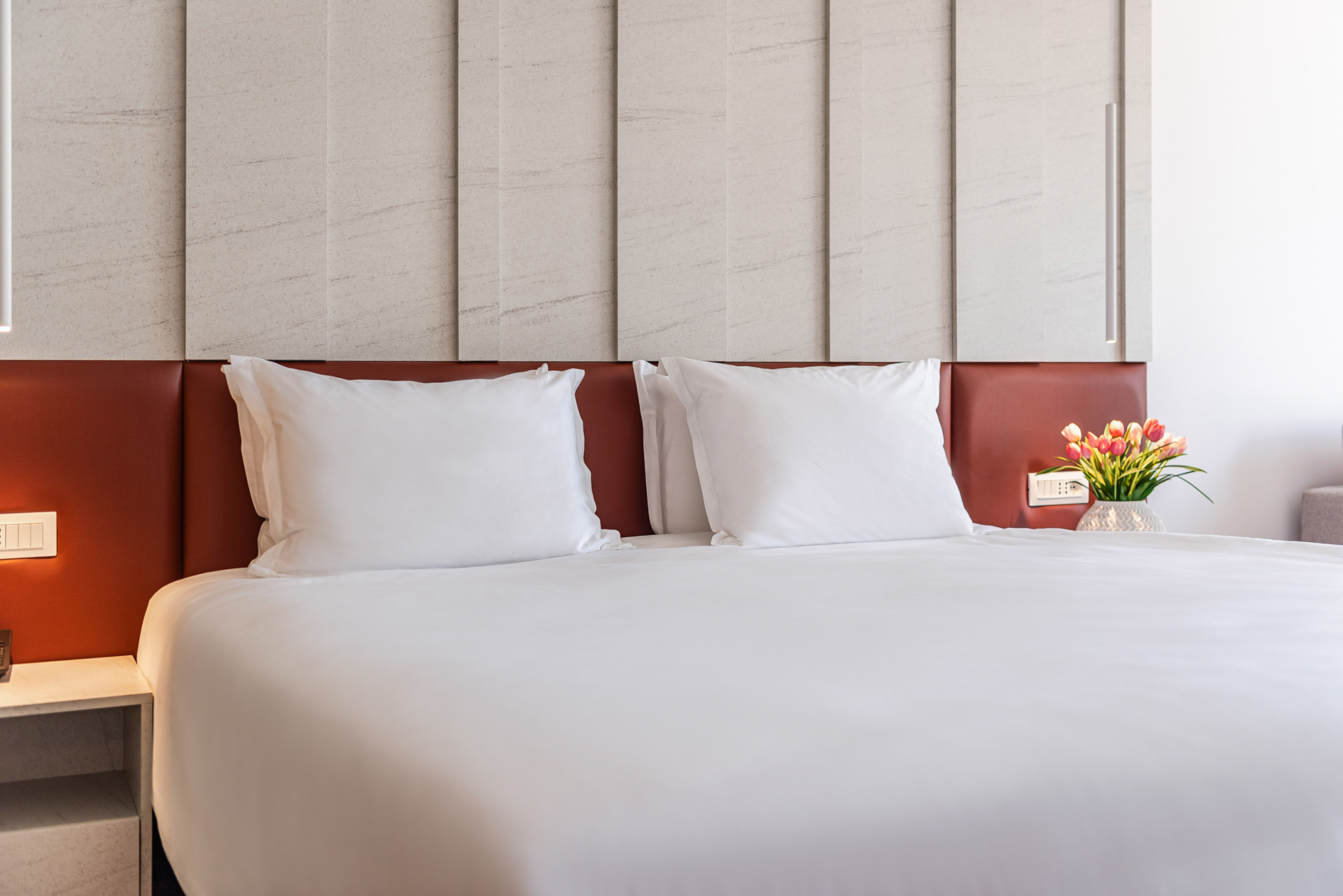 Clean white hotel bed, brown headboard, textured wall, and tulips on nightstand.