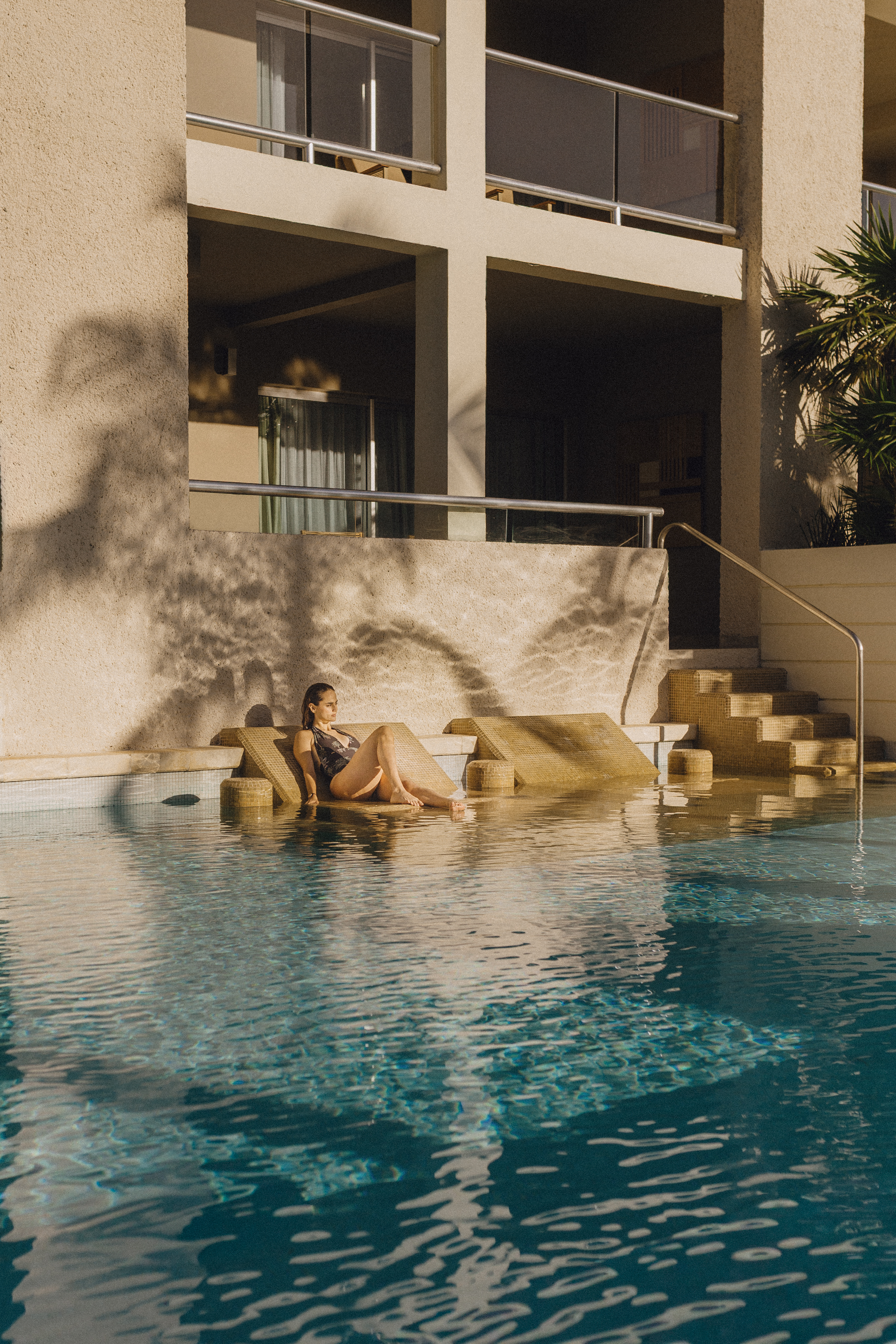 Woman in swimsuit relaxing on in-pool sunbed at sunny resort, building in background.