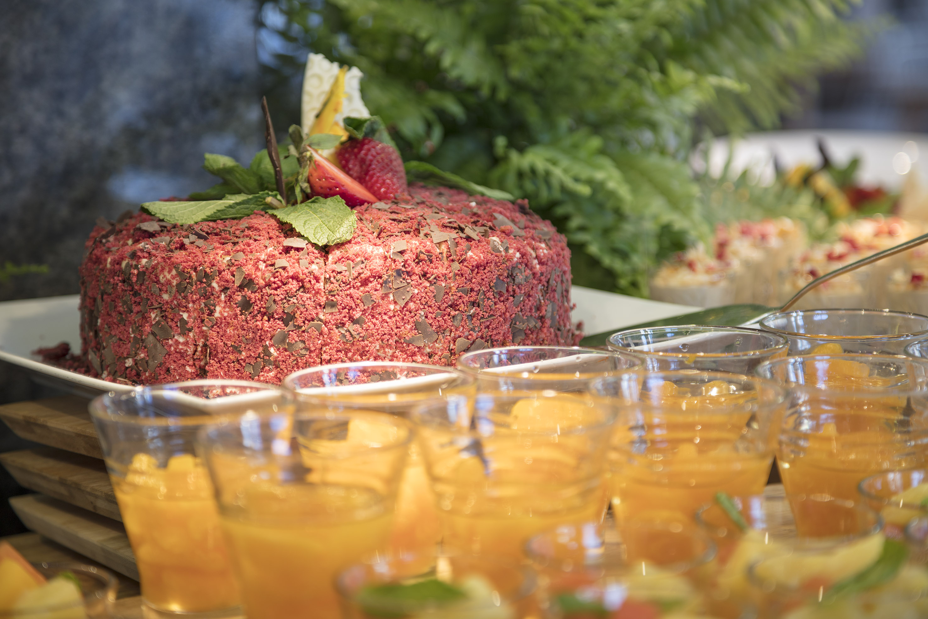 Buffet dessert table with red cake, strawberry, chocolate, and orange juice shots.