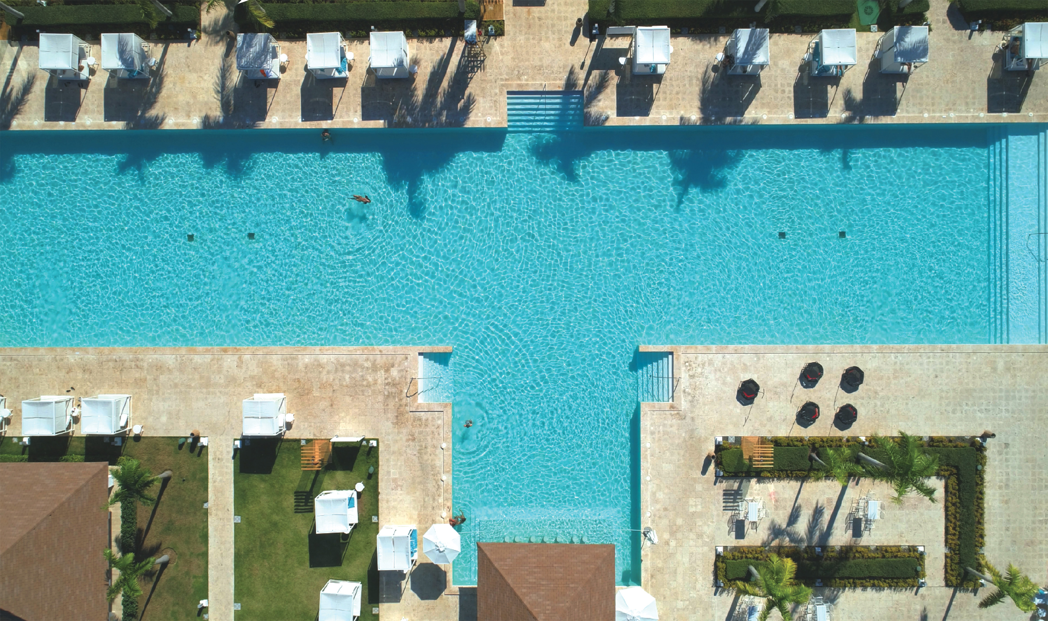 Aerial view of a T-shaped resort pool, with swimmers, cabanas, and palm trees.