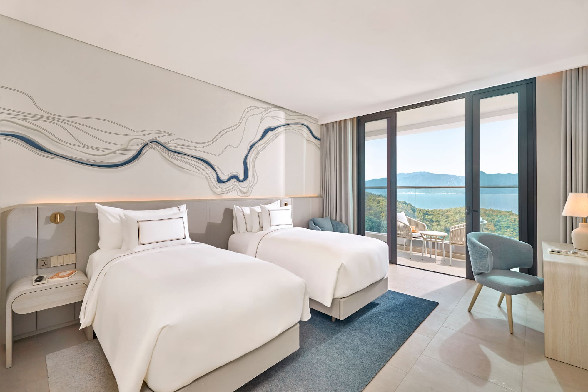 Modern hotel room with twin beds, balcony, and ocean mountain views.