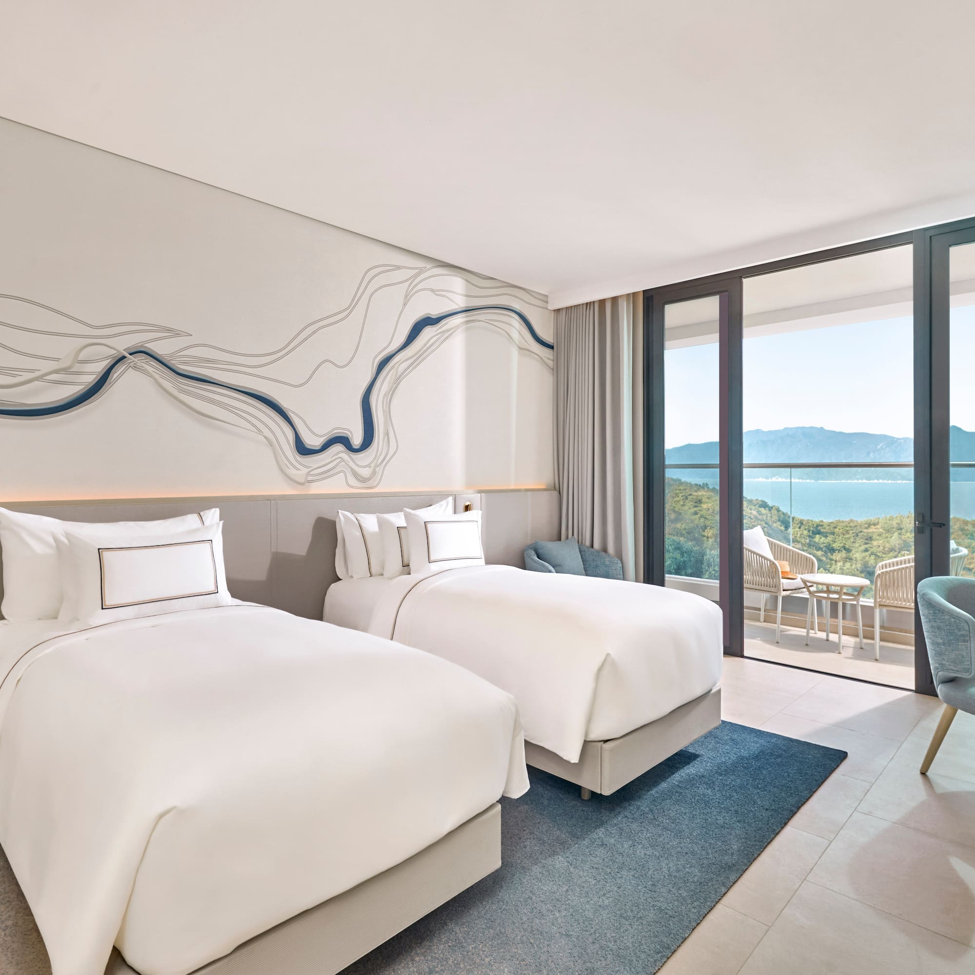 Modern hotel room with twin beds, balcony, and ocean mountain views.