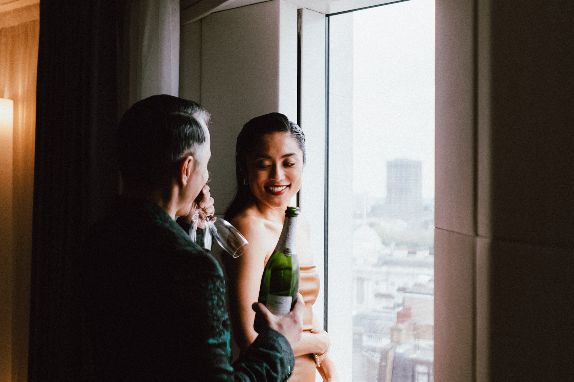 A man is holding a bottle of wine while a woman stands in front of a window.