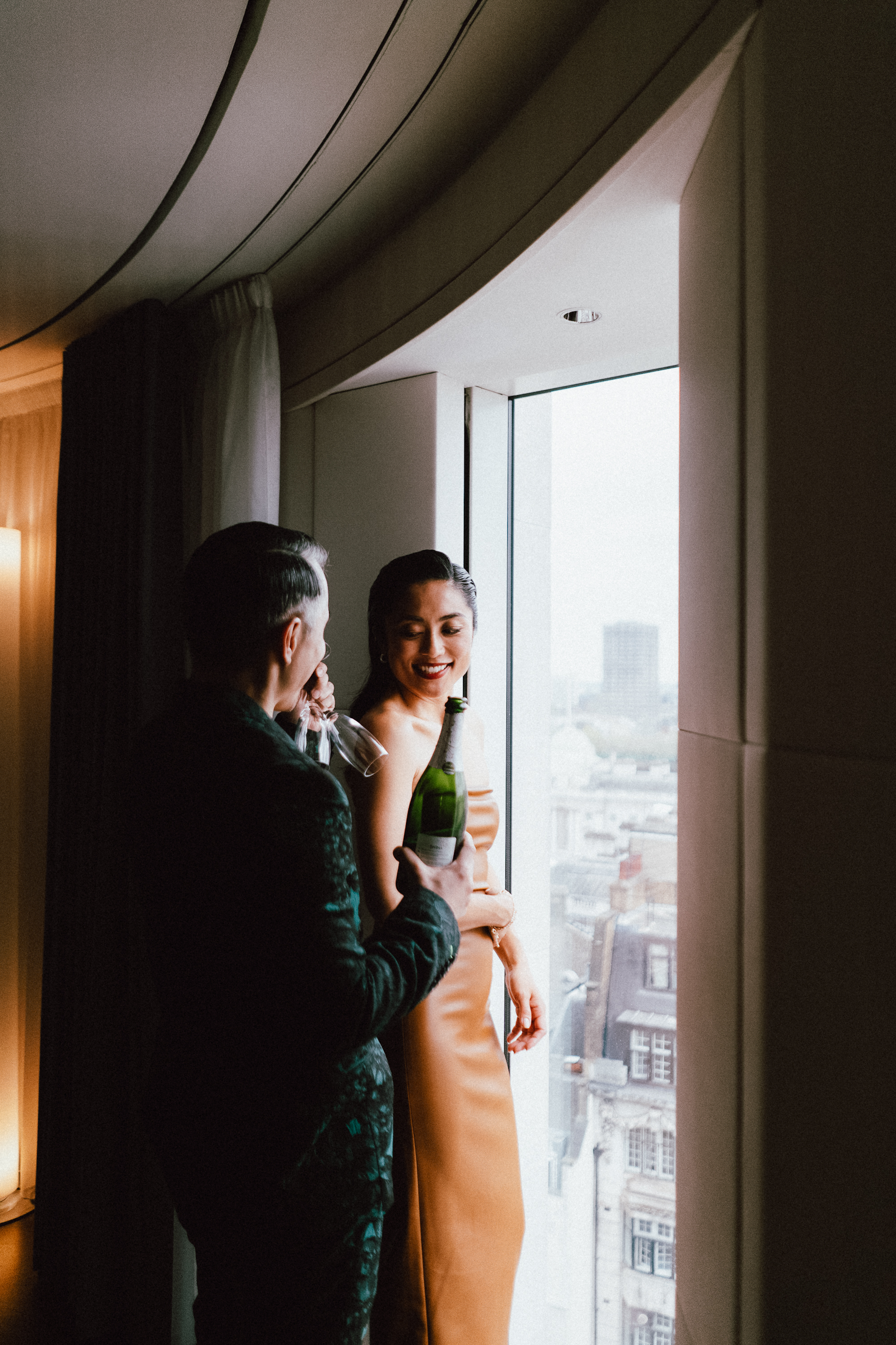 Couple holding champagne bottle and glass by window, city view.
