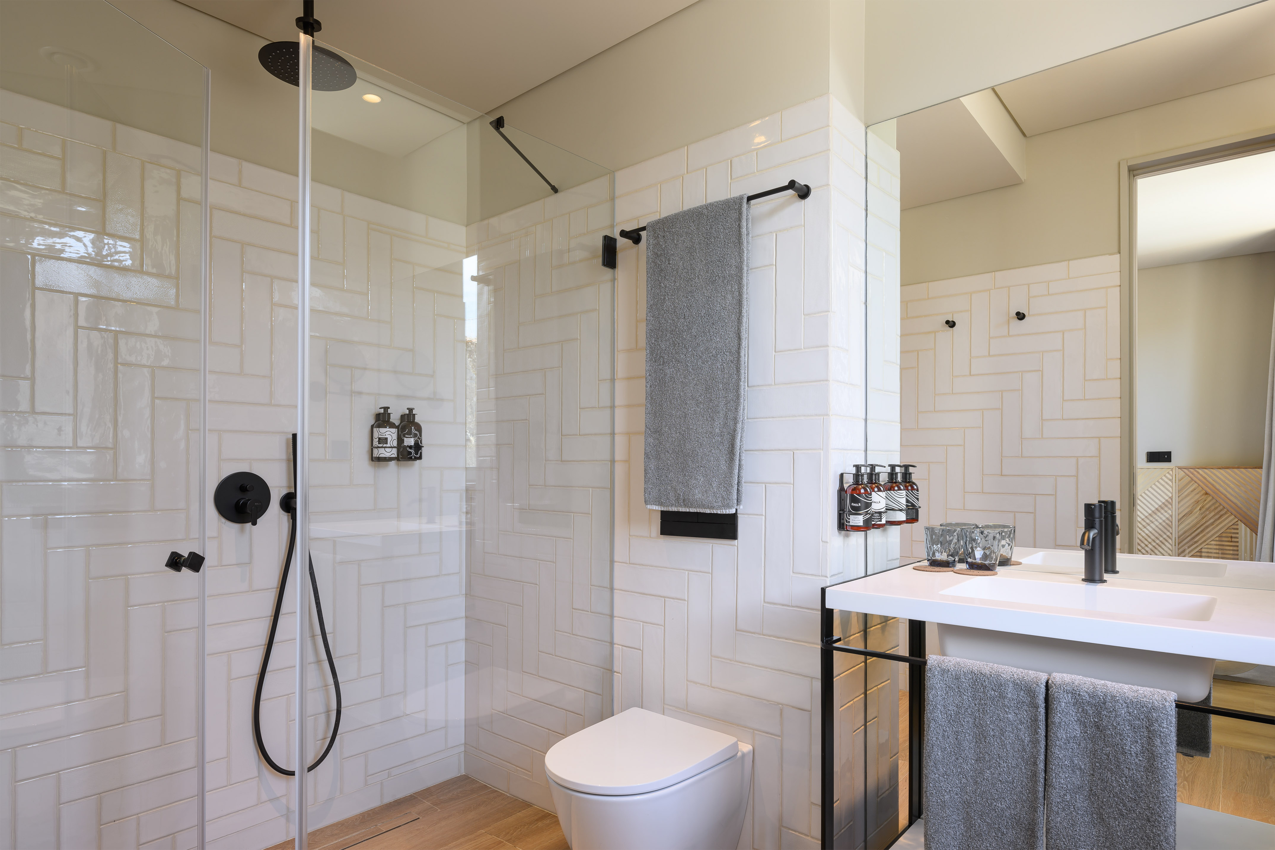Modern bathroom with white herringbone tiles, glass shower, black fixtures, toilet, and vanity.