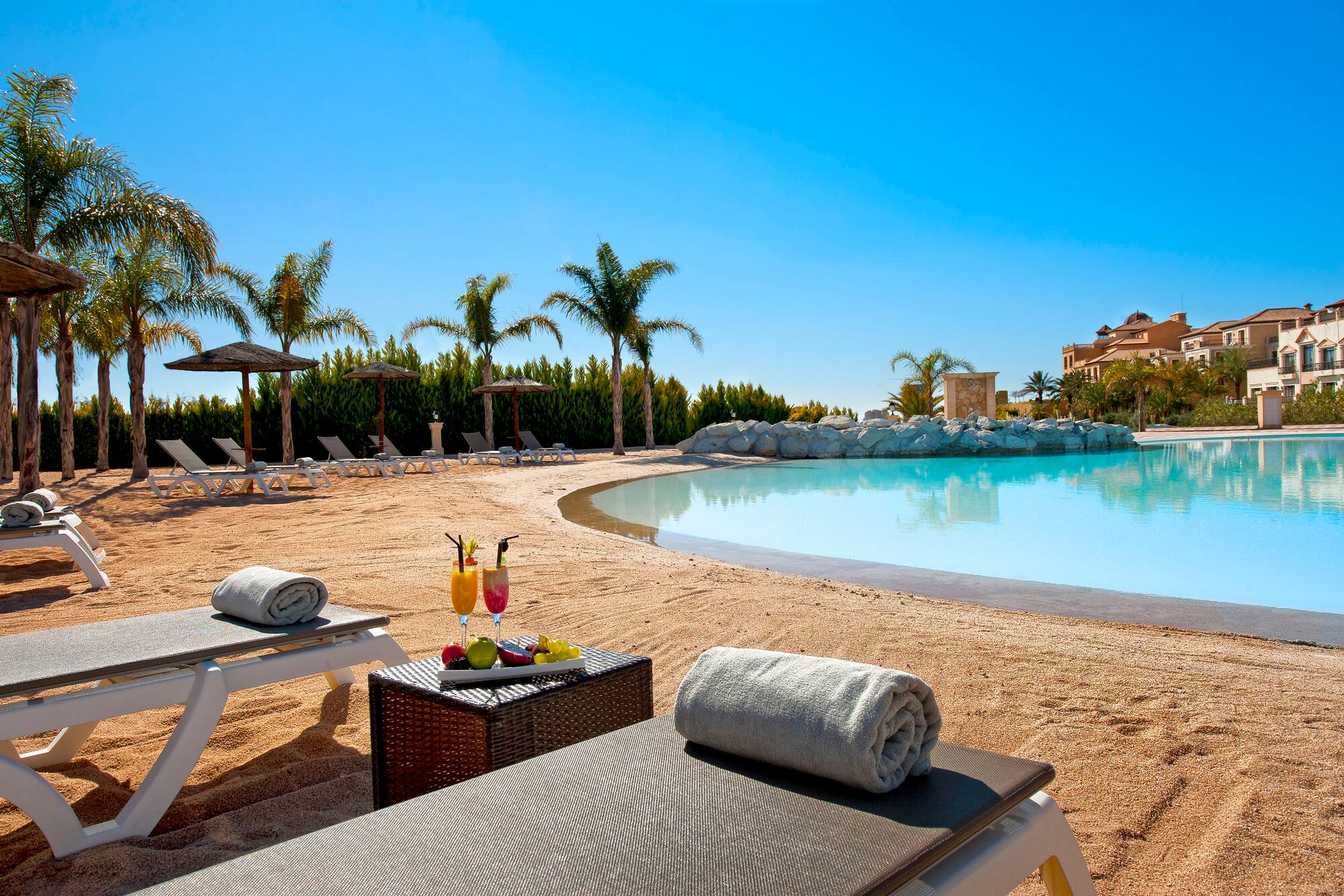 Sunny resort pool with sandy beach, palm trees, loungers, and cocktails.