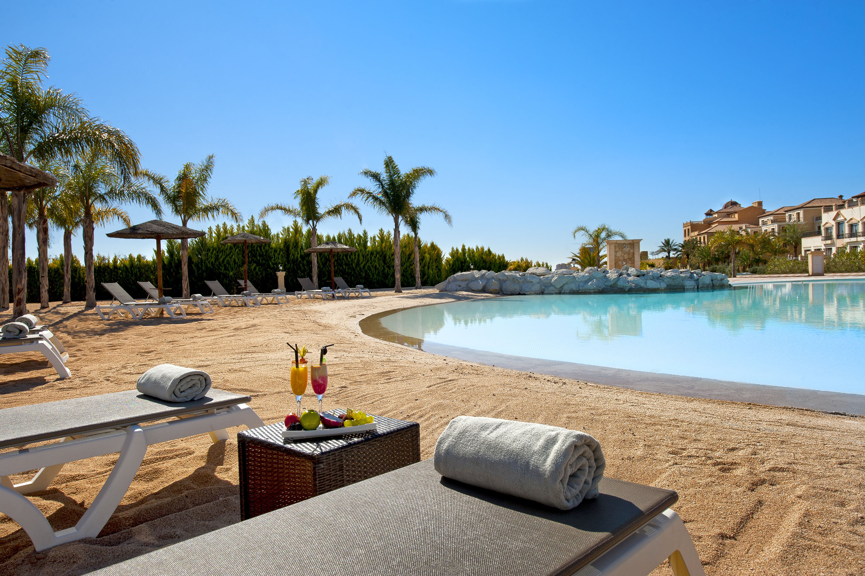 Sunny resort pool with sandy beach, palm trees, loungers, and cocktails.
