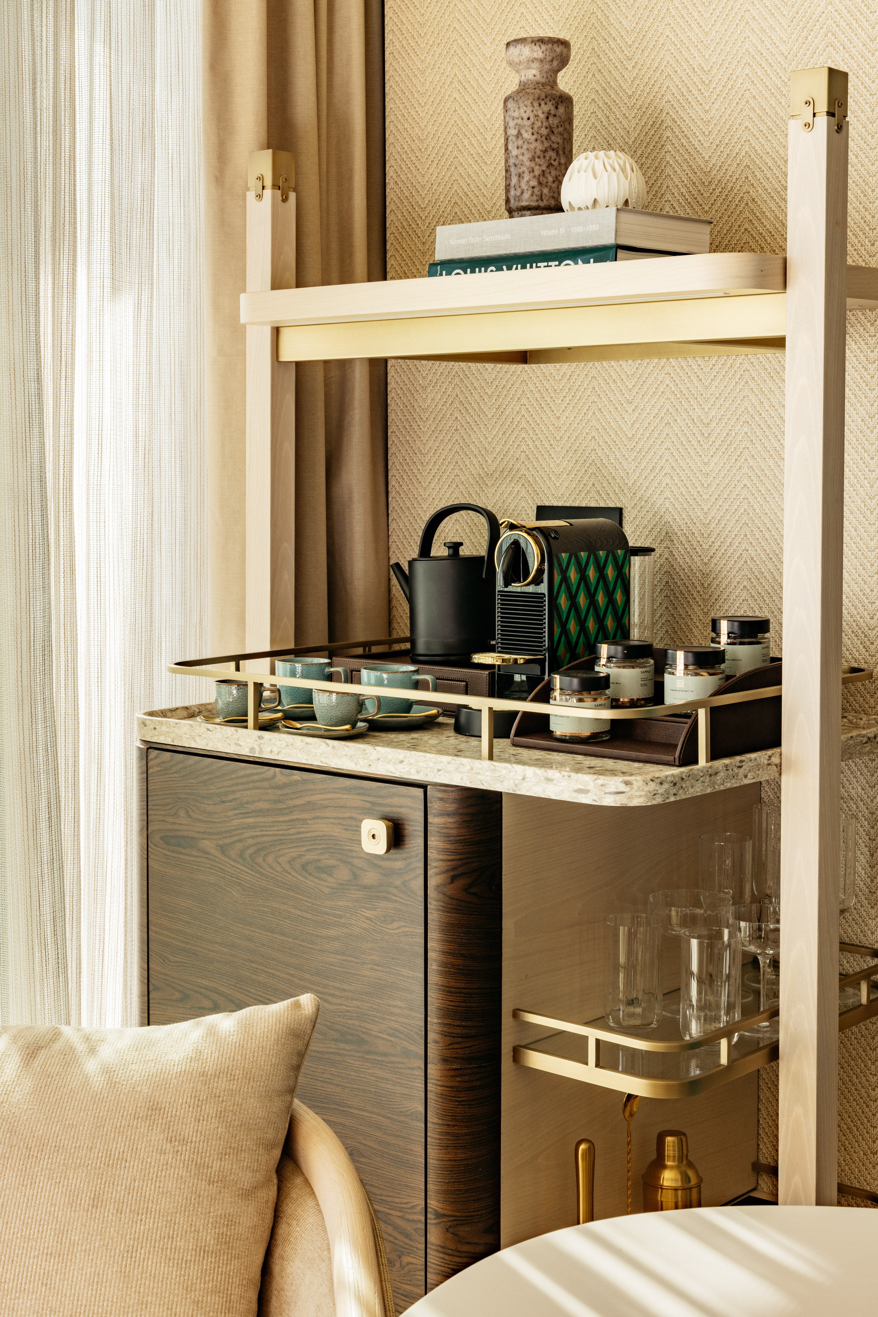 Modern hotel minibar with Nespresso, tea set, glassware, and books on shelves.