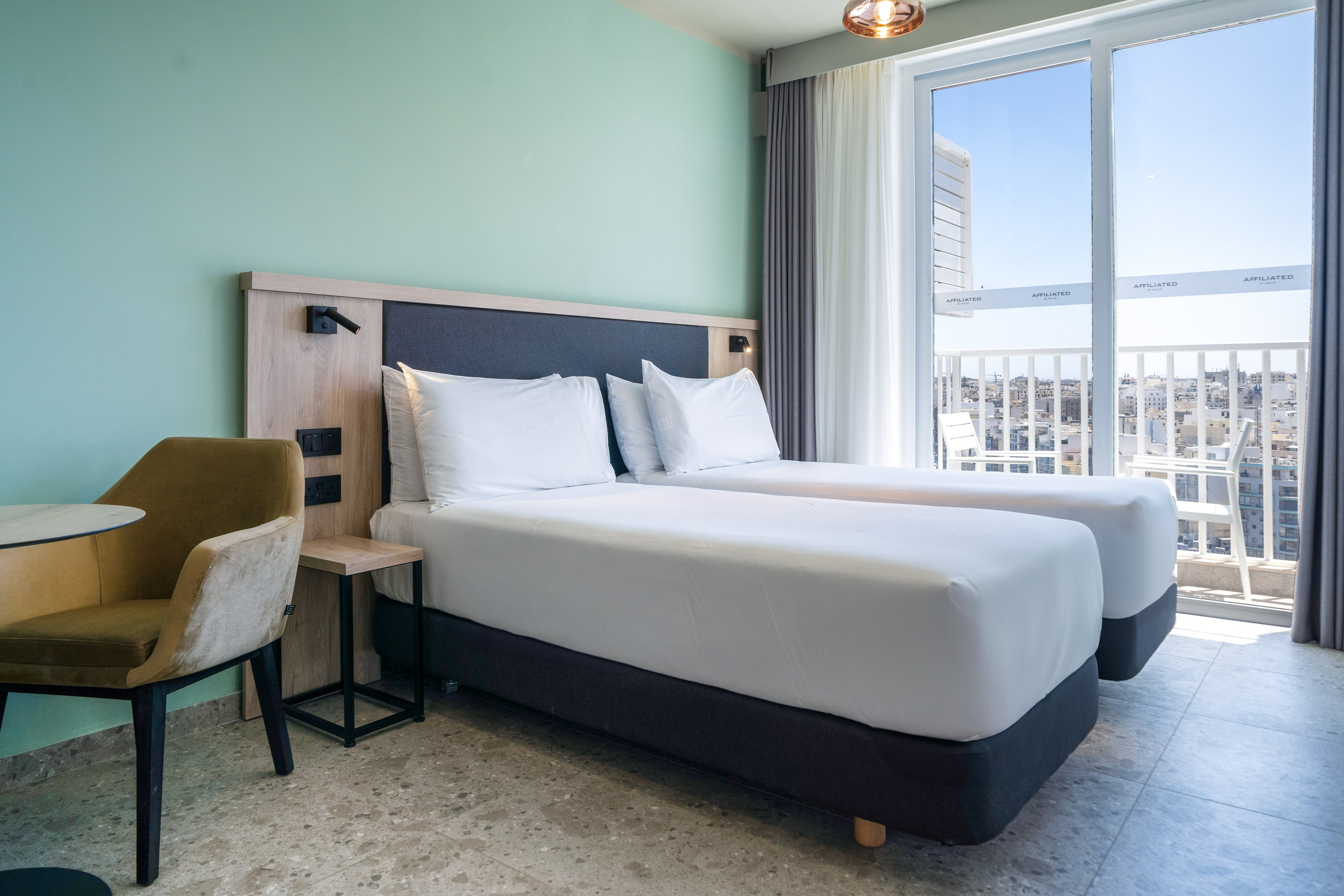 Modern hotel room with two twin beds, balcony city view, and green wall.