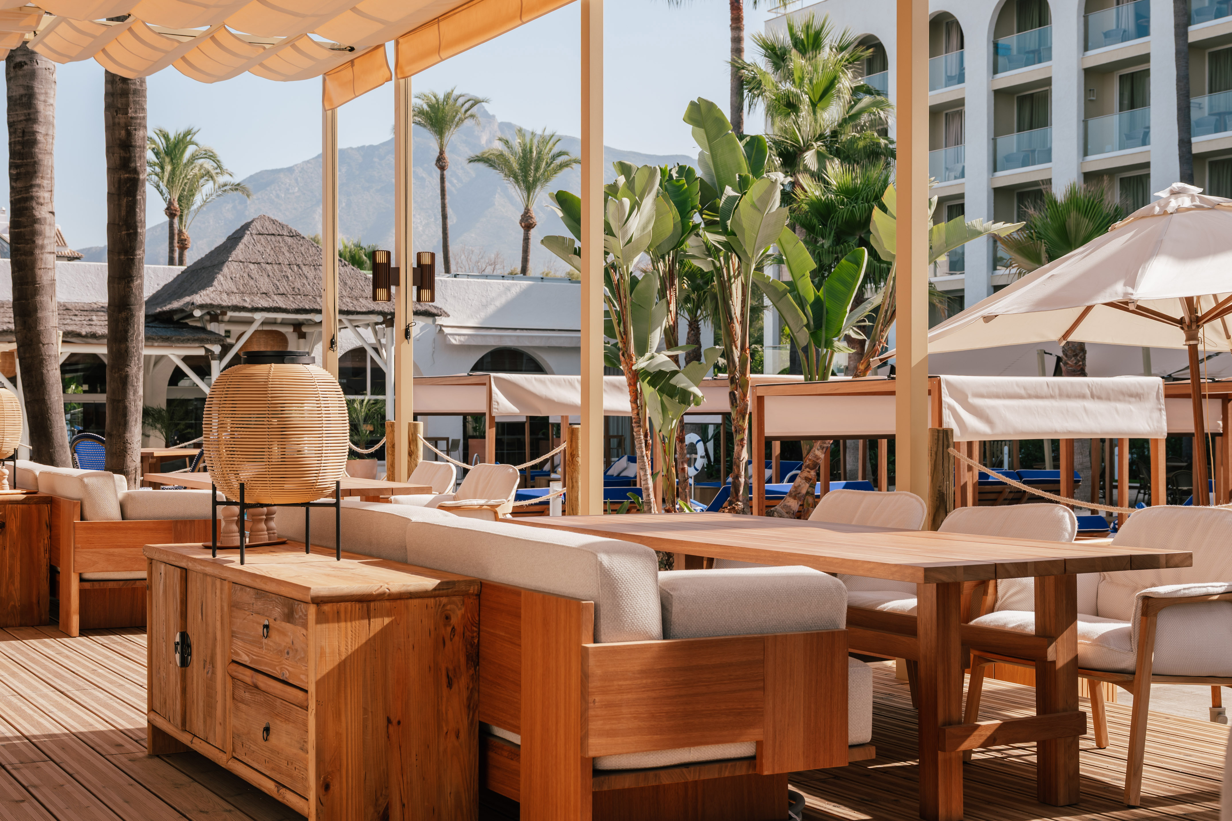 Outdoor resort patio with wooden furniture, tropical plants, and mountains.