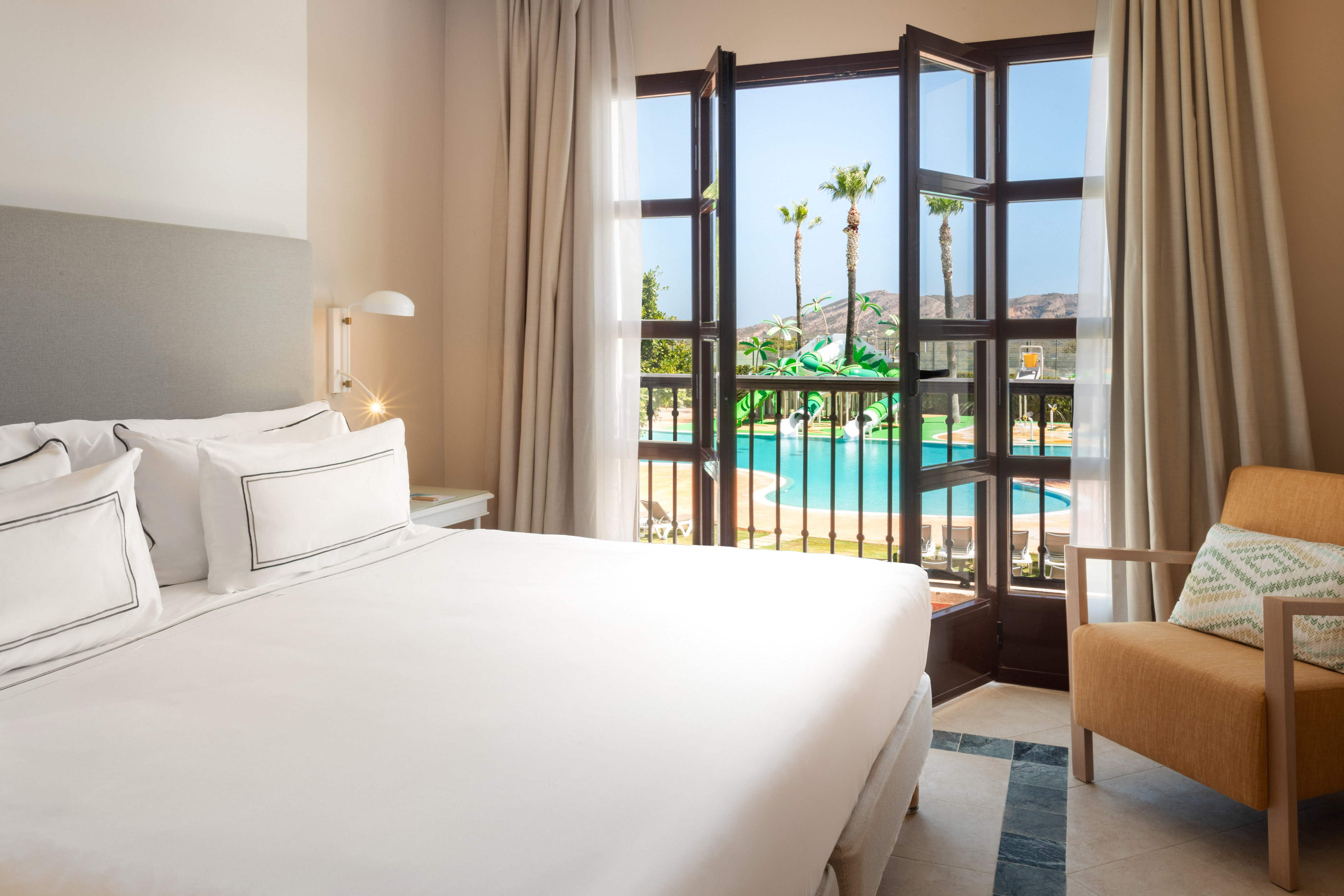 Resort bedroom with pool view, waterslides, and palm trees from open window.