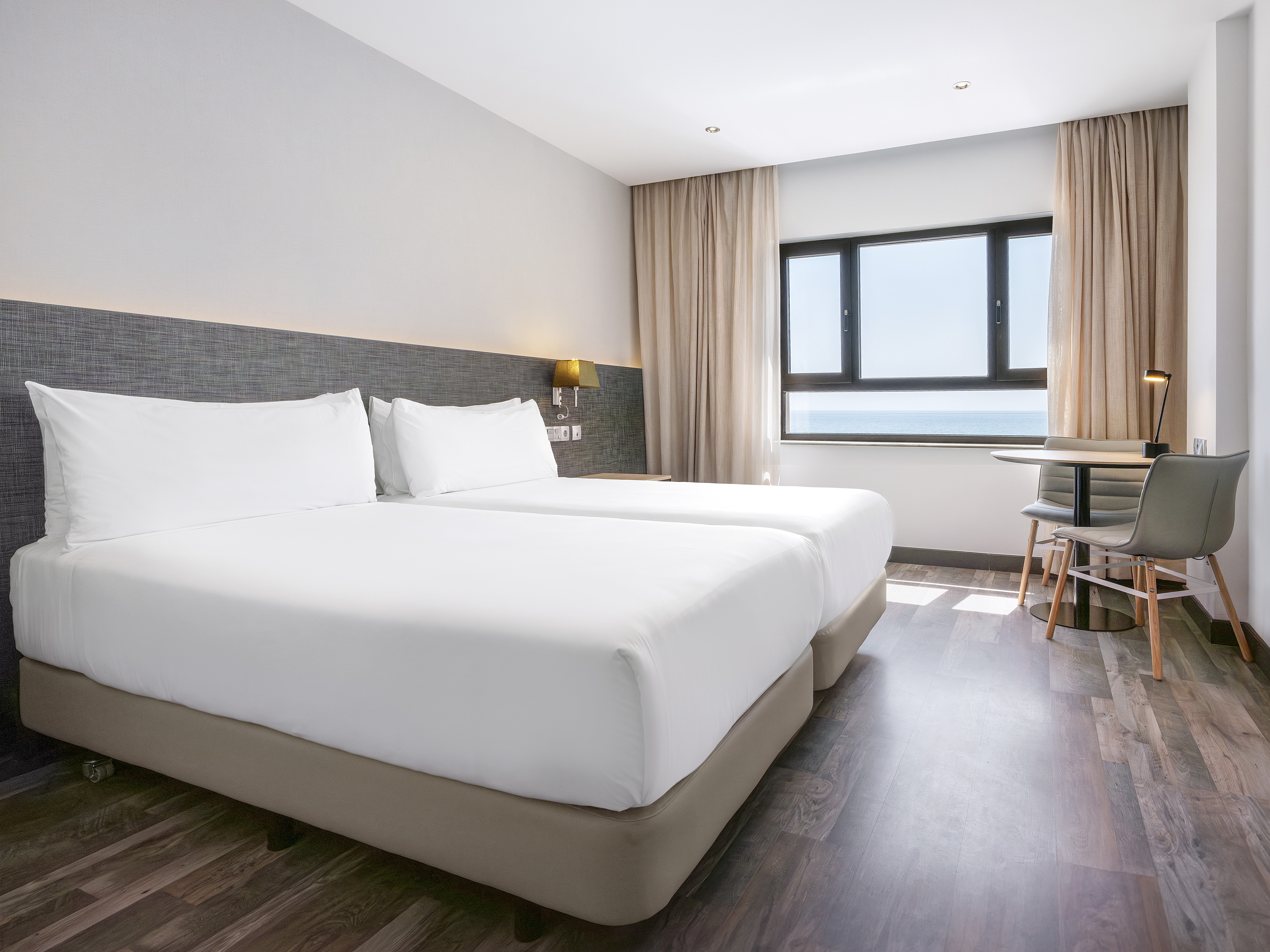 Modern hotel twin room with ocean view, white beds, and dark wood floor.