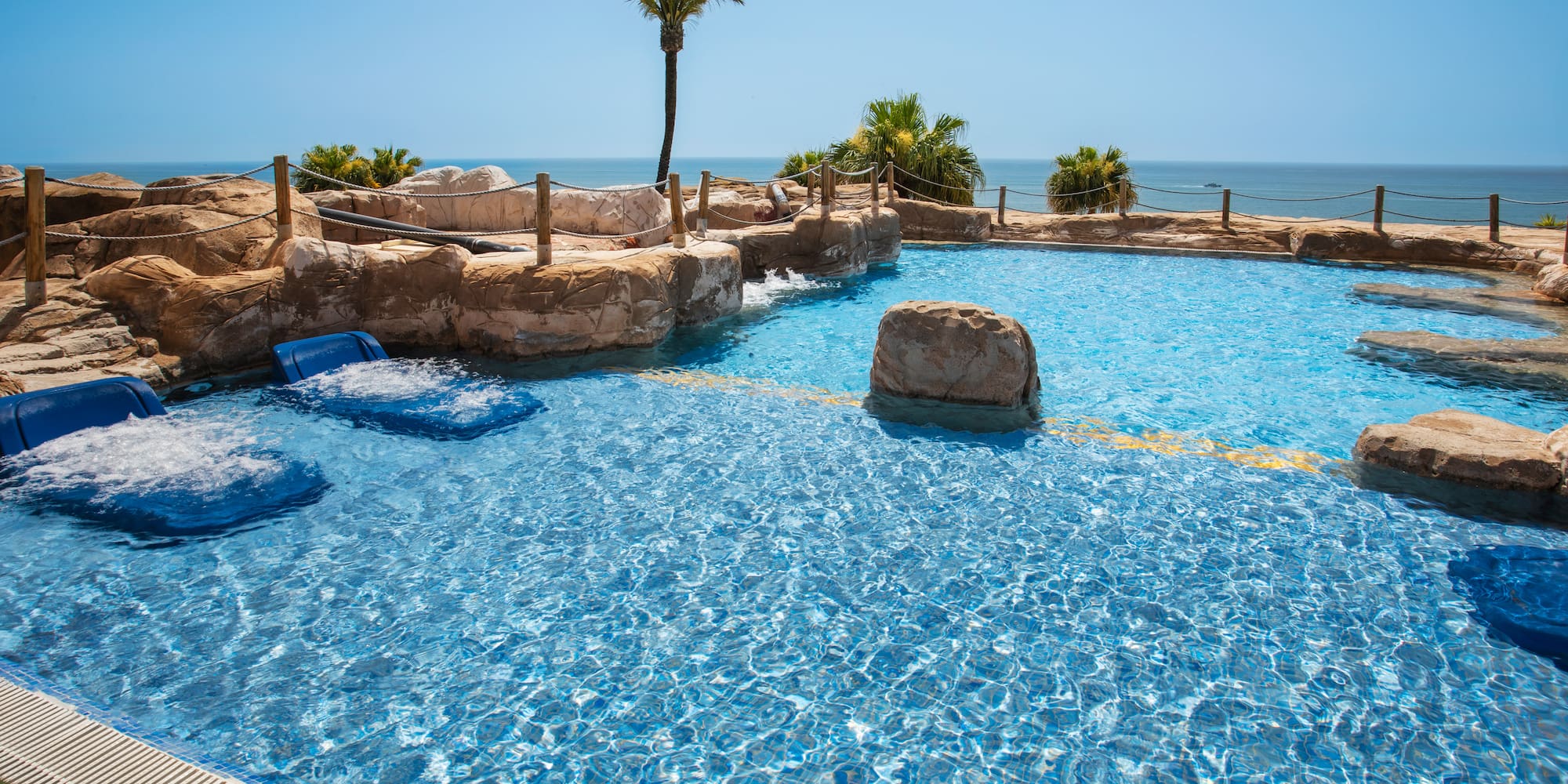 Hotel swimming pool with rock features, spa jets, palm trees, and ocean view.