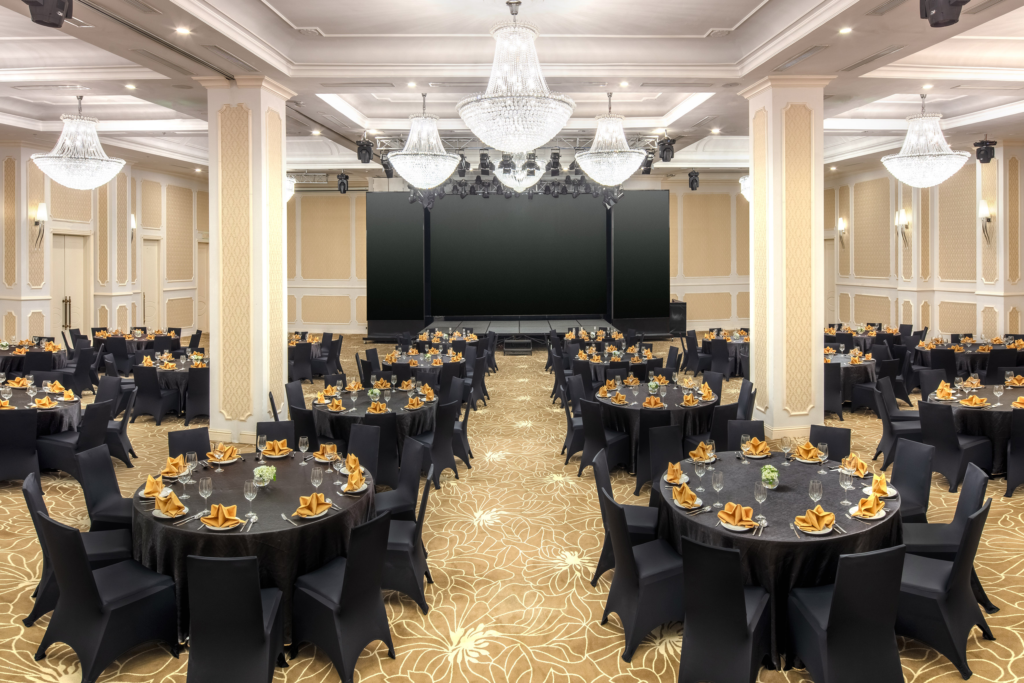 Elegant ballroom set with black banquet tables, chairs, stage, and crystal chandeliers.