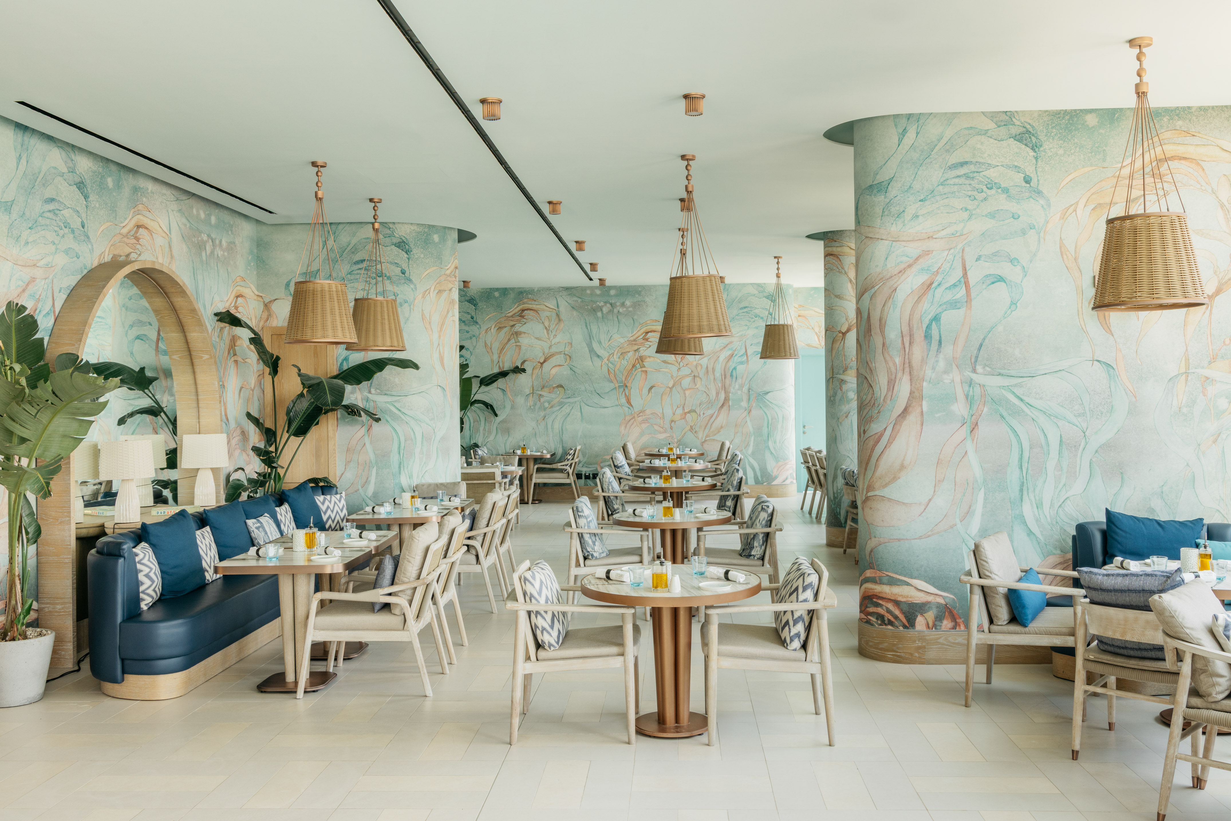 Tropical restaurant with botanical wallpaper, blue seating, and wicker lights.